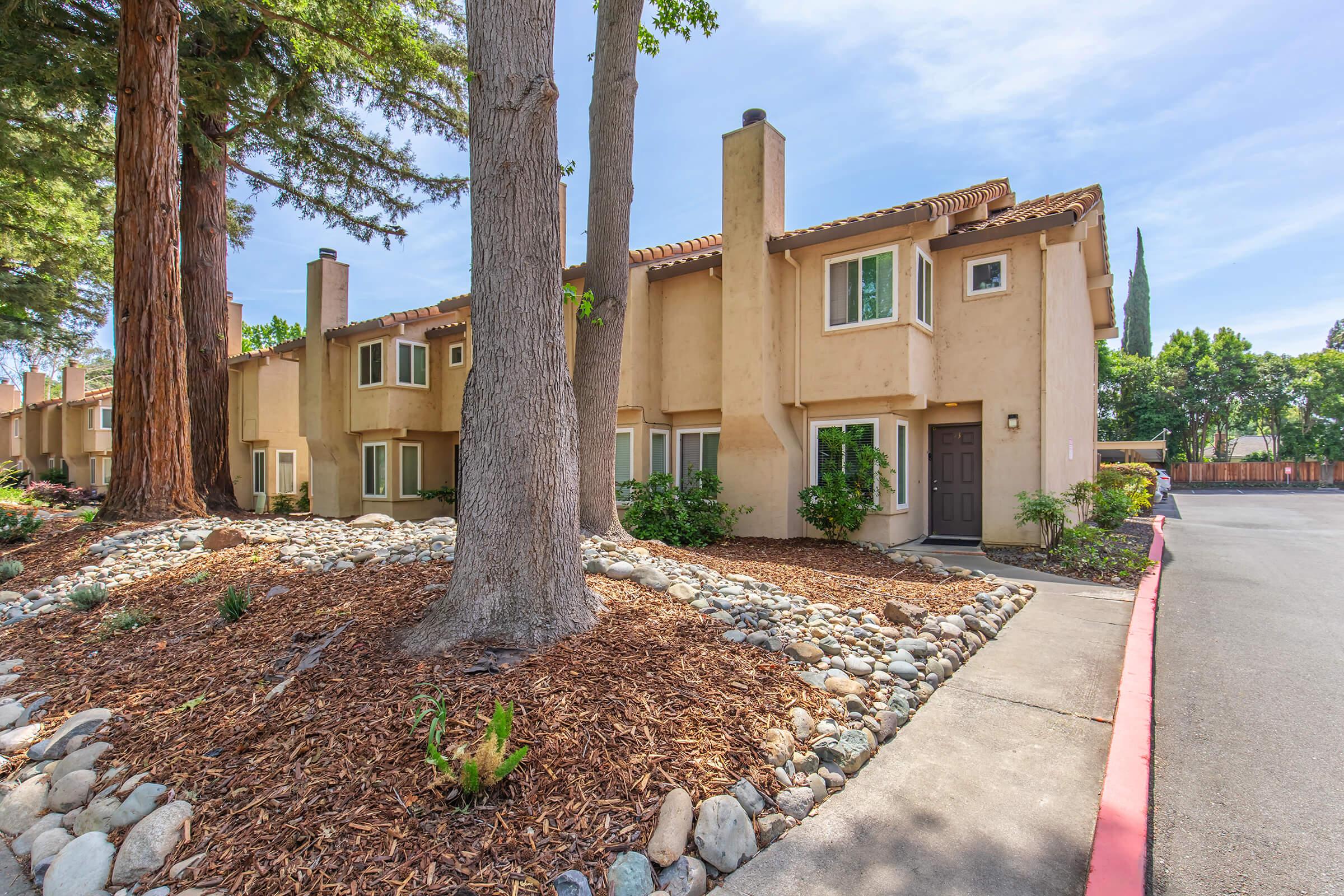 A view of a residential building with a light brown exterior, surrounded by trees and landscaping. The front features a pathway, decorative rocks, and small plants. The scene is bright with a clear blue sky, showcasing a peaceful and pleasant neighborhood atmosphere.