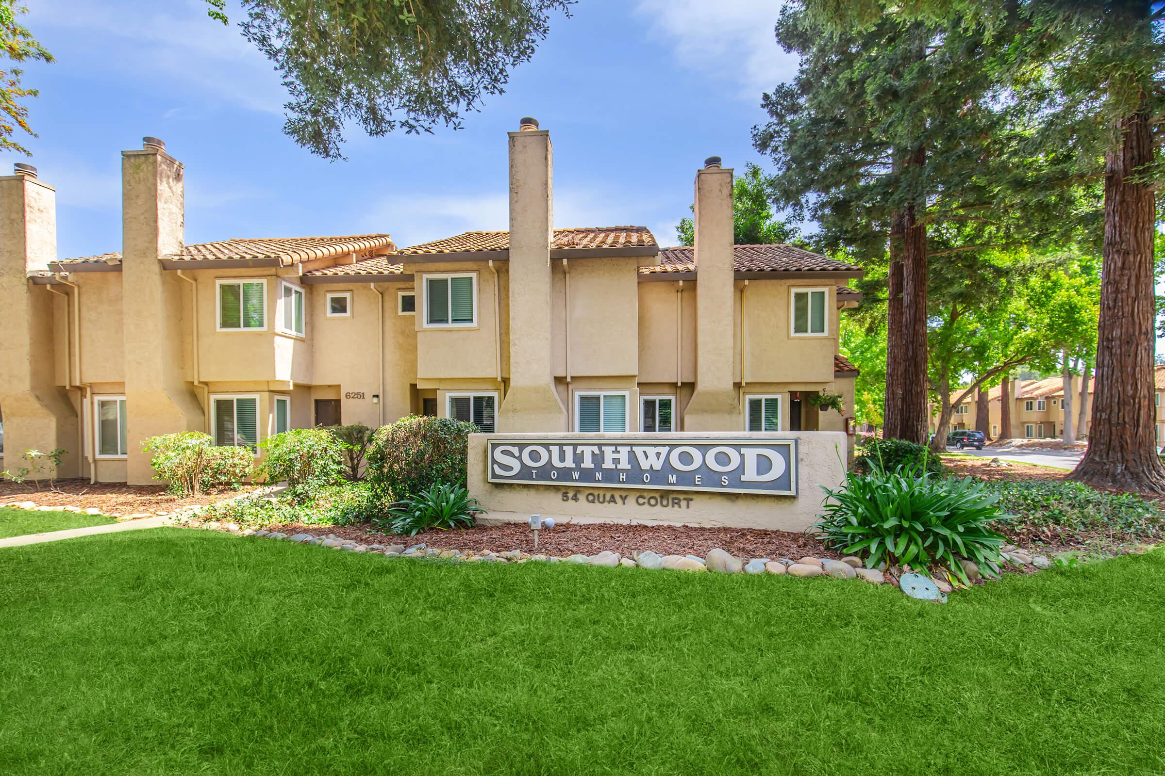 A residential complex named "Southwood Townhouses" featuring a prominent sign at the entrance. The setting includes well-maintained landscaping with green grass and shrubs, along with a clear view of the townhouses under a bright blue sky and tall trees.