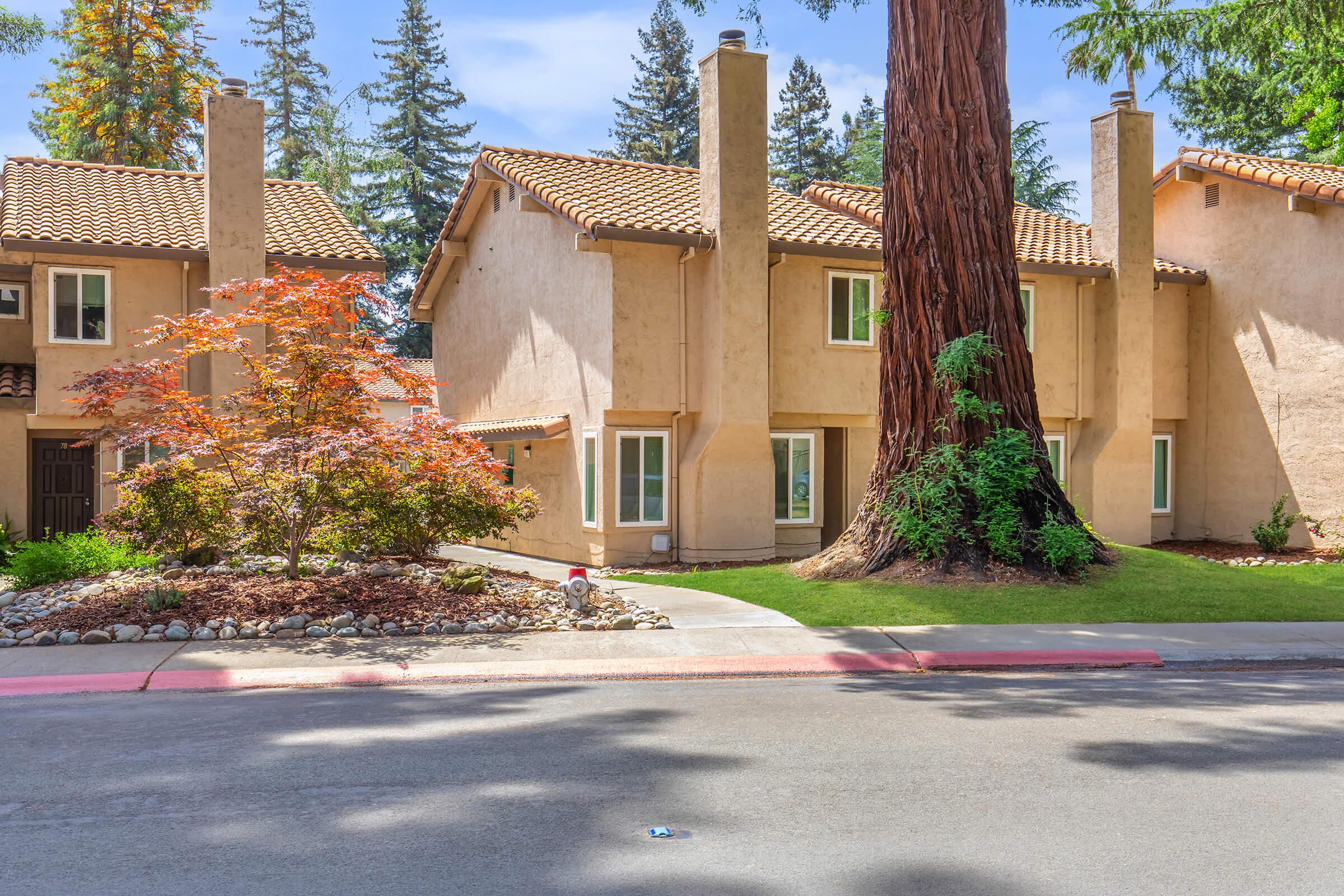 A quaint residential area features two-story buildings with stucco exteriors and tiled roofs. Lush green trees surround the homes, and colorful landscaping adds charm to the scene. A smooth road leads to the entrance of the properties, enhancing the inviting atmosphere of the neighborhood.