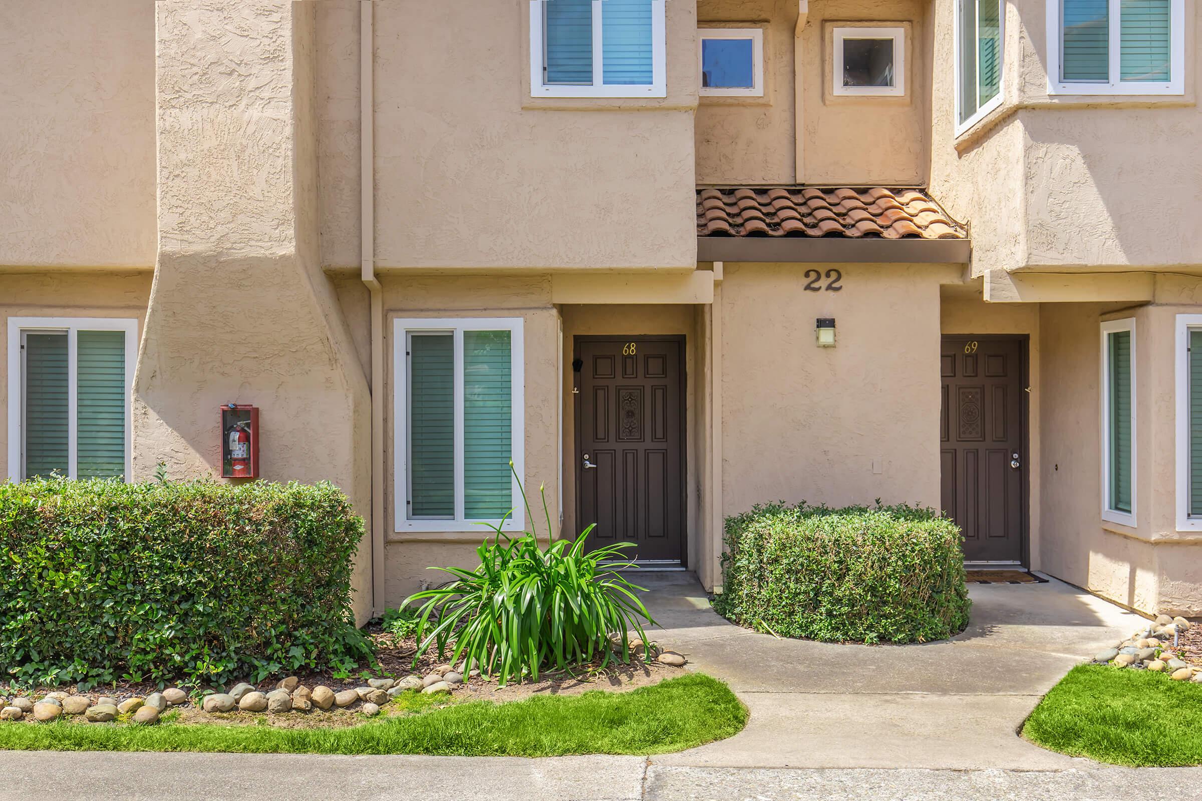 Two residential doors situated in a light-colored building with a tile roof. The left door is labeled "22" and the right door labeled "68." Each door has windows on either side, and there are well-maintained shrubs and grass in front of the entrance. The scene is bright and inviting.