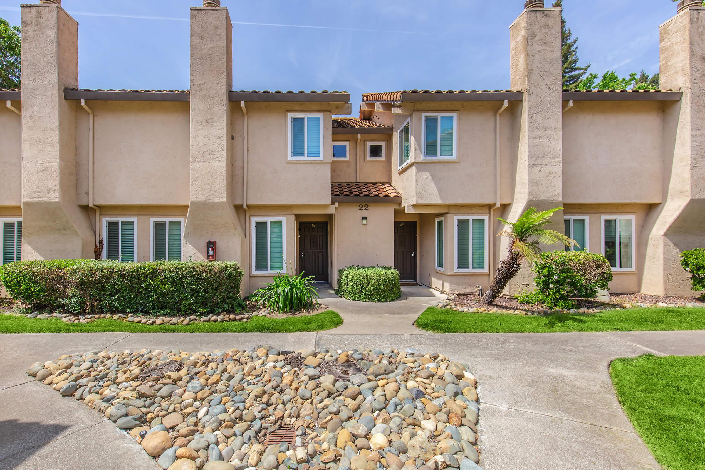 A two-story townhouse complex with light-colored stucco walls and brown roofs. The entrance features two doors, bordered by small shrubs and landscaping. A rocky pathway leads to the entrance, and the area is well-maintained with a lawn and palm trees. Blue skies are visible in the background.