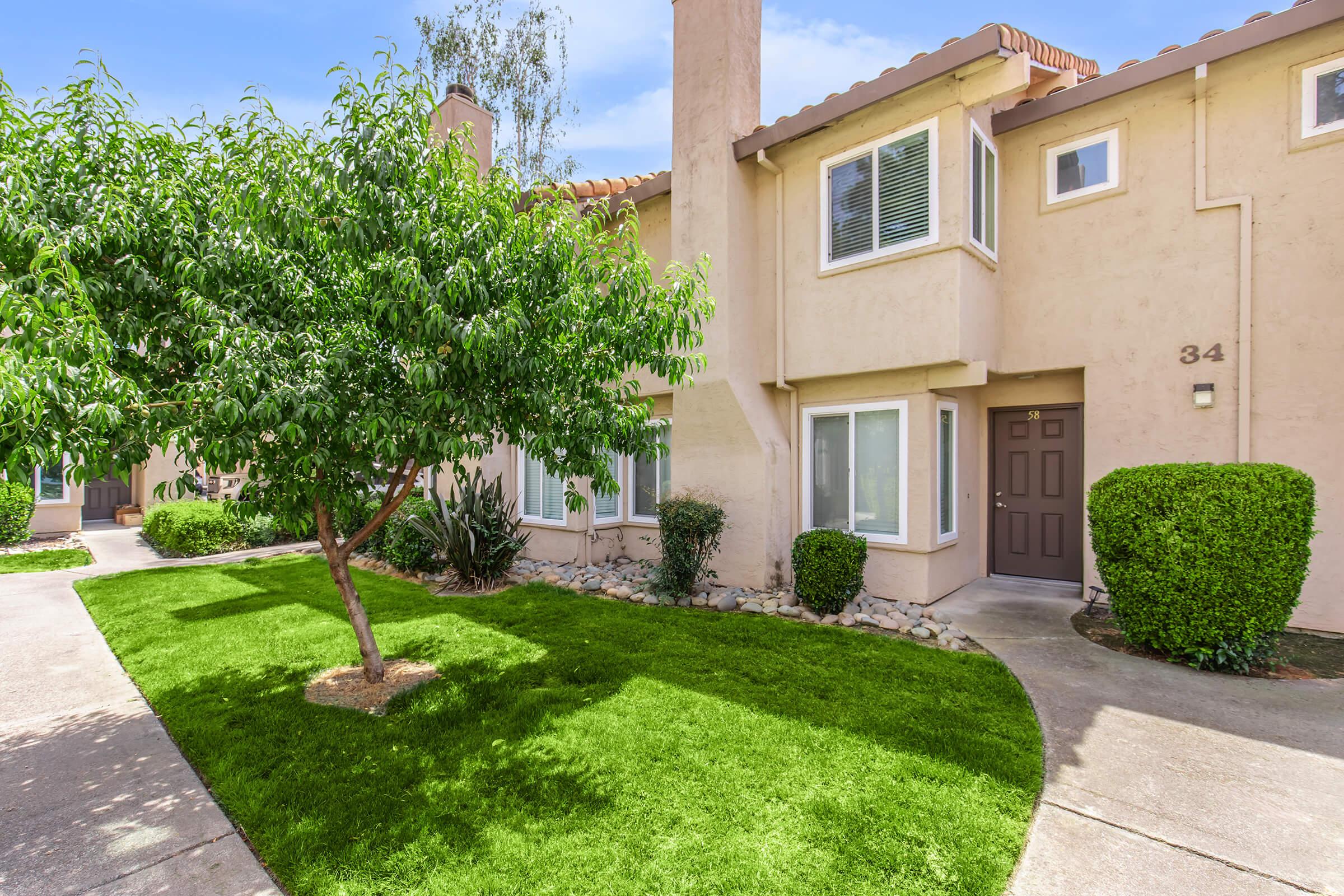 A well-maintained townhouse complex featuring a green lawn, a small tree, and path leading to a front door. The building has two stories with beige stucco walls, and several windows allowing natural light. The area is clean and includes shrubs and rocks for landscaping.