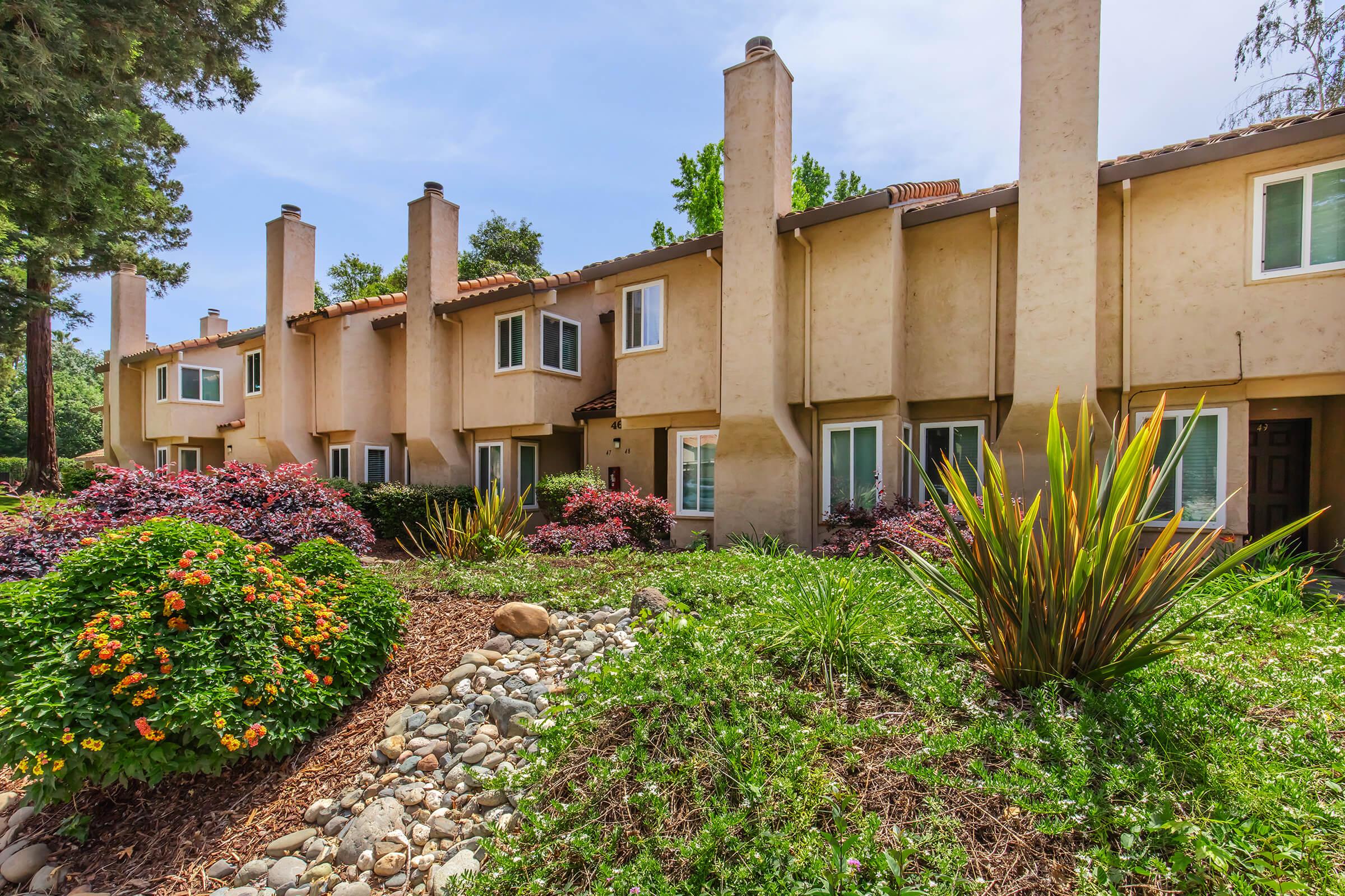 A row of beige townhouses with multiple windows, surrounded by vibrant flower beds and greenery. The landscape features colorful plants and a pebbled area, under a clear blue sky with a few trees in the background, creating a welcoming and serene environment.