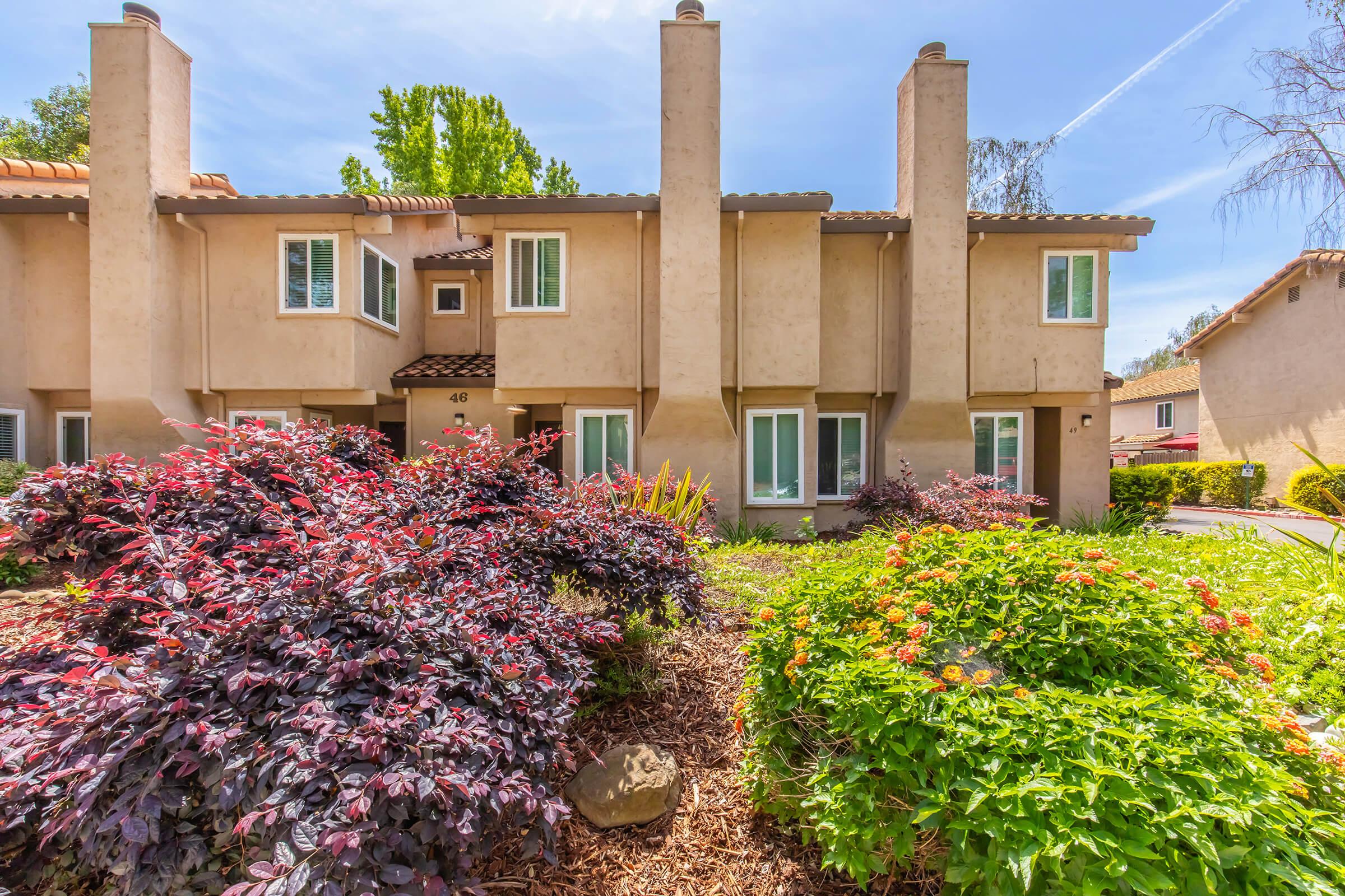 View of a two-story residential building featuring a beige exterior with multiple windows, surrounded by colorful landscaping. The foreground includes lush, vibrant shrubs and flowers, while the background showcases a clear blue sky and trees, creating a pleasant and inviting atmosphere.