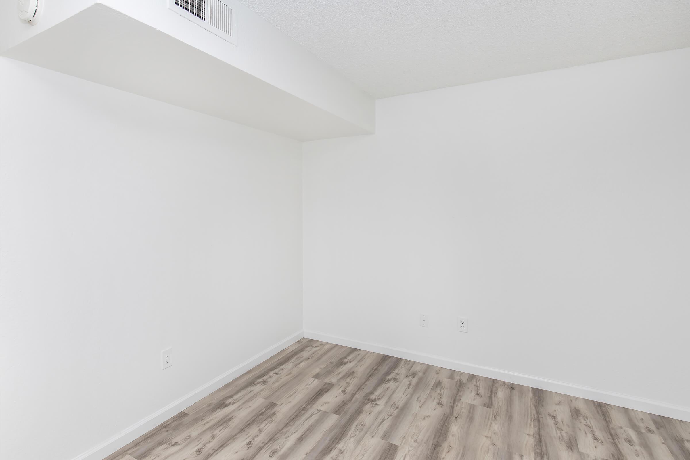 Empty interior of a room with white walls and light-colored laminate flooring. There are no furniture or decorations, creating a minimalist and spacious appearance. A small air vent is visible on the wall near the ceiling.