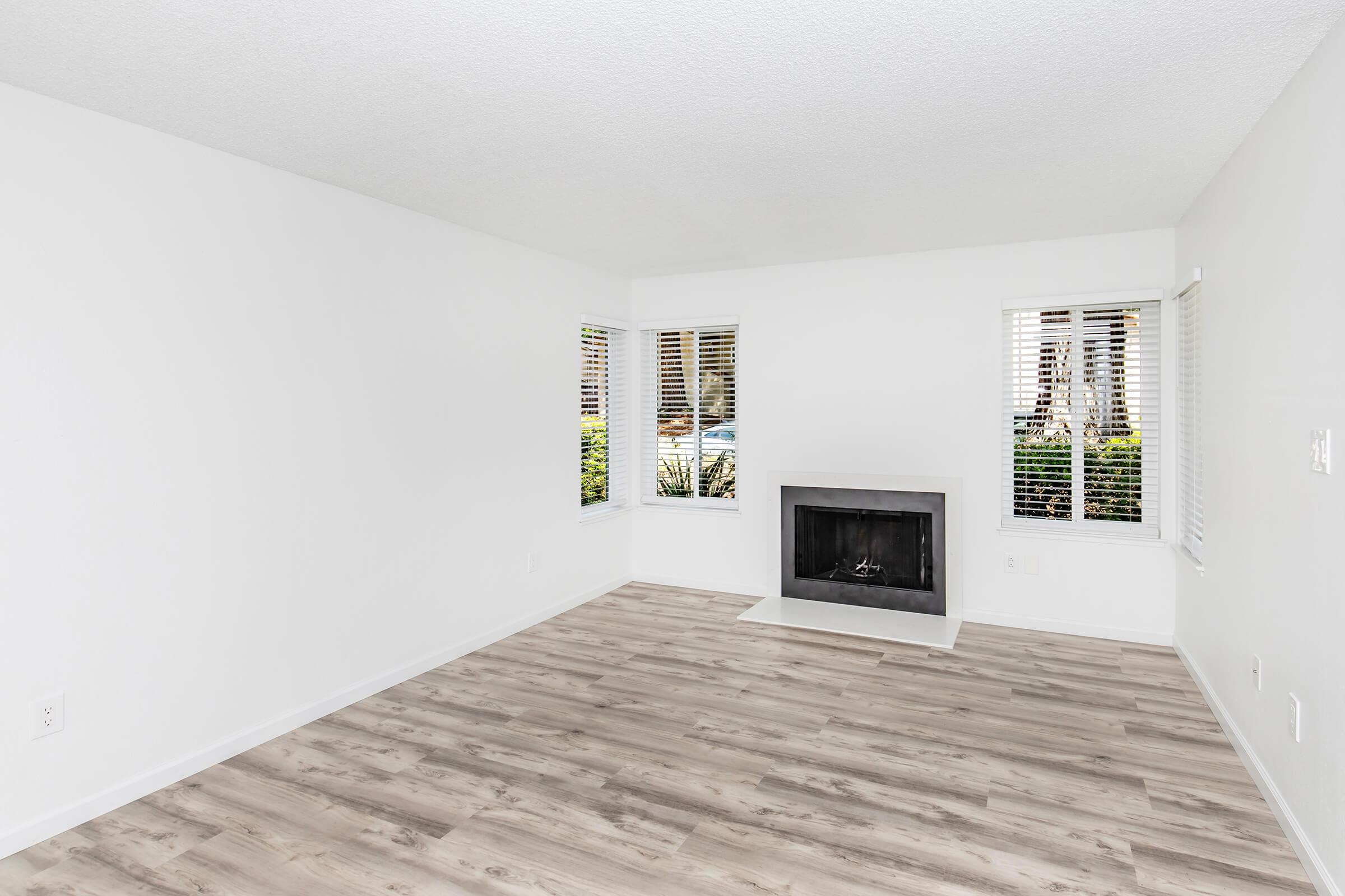 A bright, empty living room with white walls and light wood flooring. It features large windows with white blinds on two walls, allowing natural light to enter. A modern fireplace is centered on one wall, and the space is minimalistic and uncluttered, providing a clean and open atmosphere.