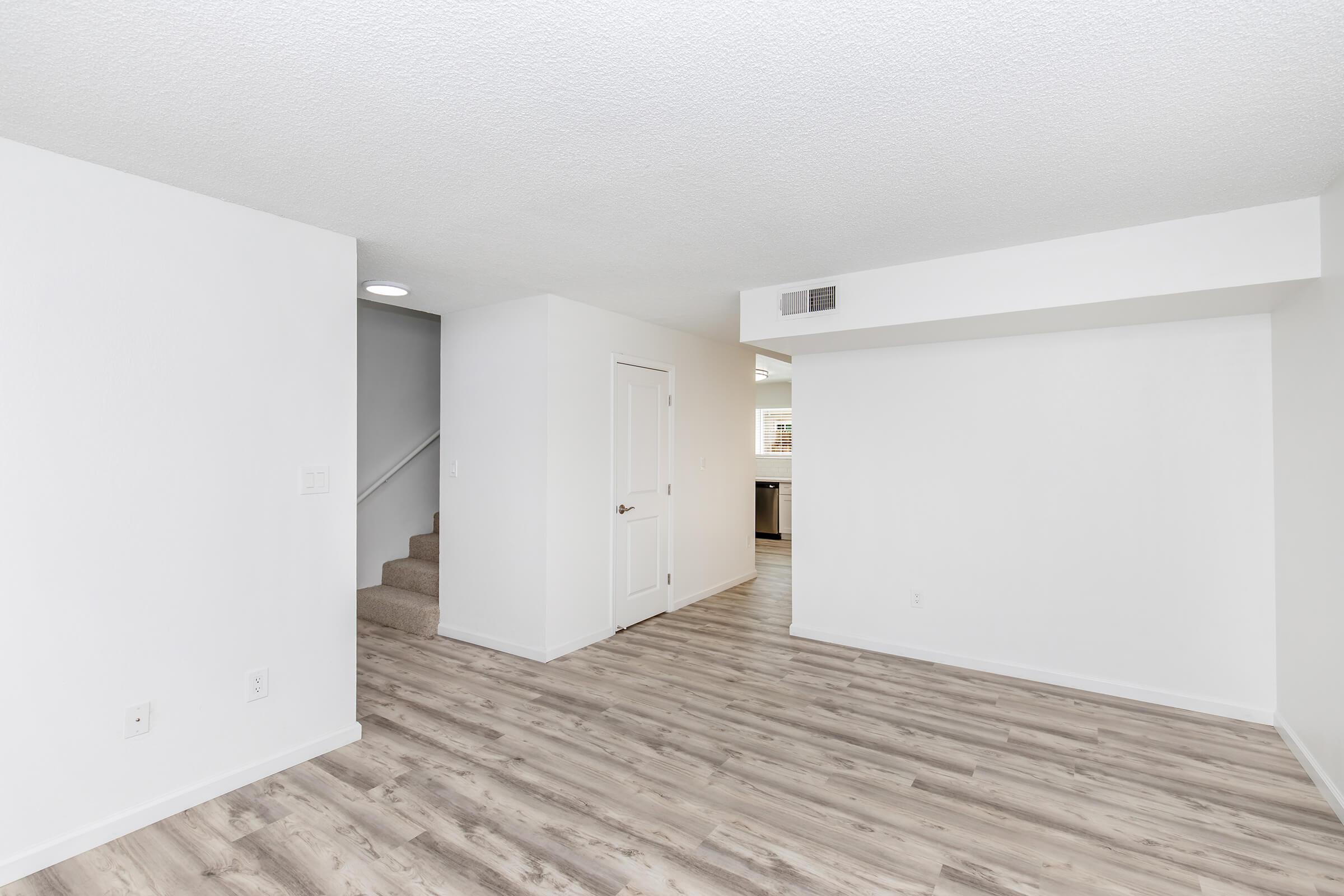 A spacious, modern living area with light-colored walls and new laminate flooring. To the left, there is a doorway leading to a staircase, and in the background, a glimpse of the kitchen area can be seen. The room is well-lit with natural light and has a clean, minimalistic design.