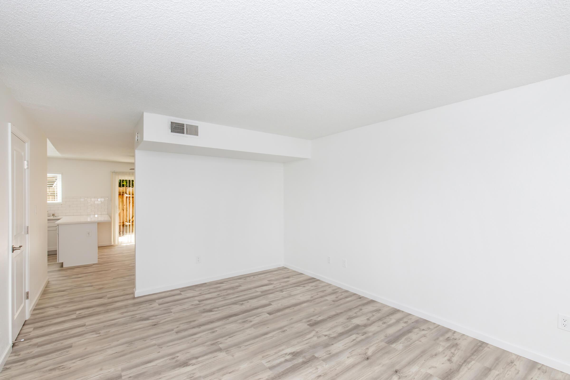 A spacious, empty room with light-colored walls and wooden laminate flooring. A doorway on the left leads to a kitchen area, and large windows allow natural light to enter. The space is clean and ready for furniture arrangement.