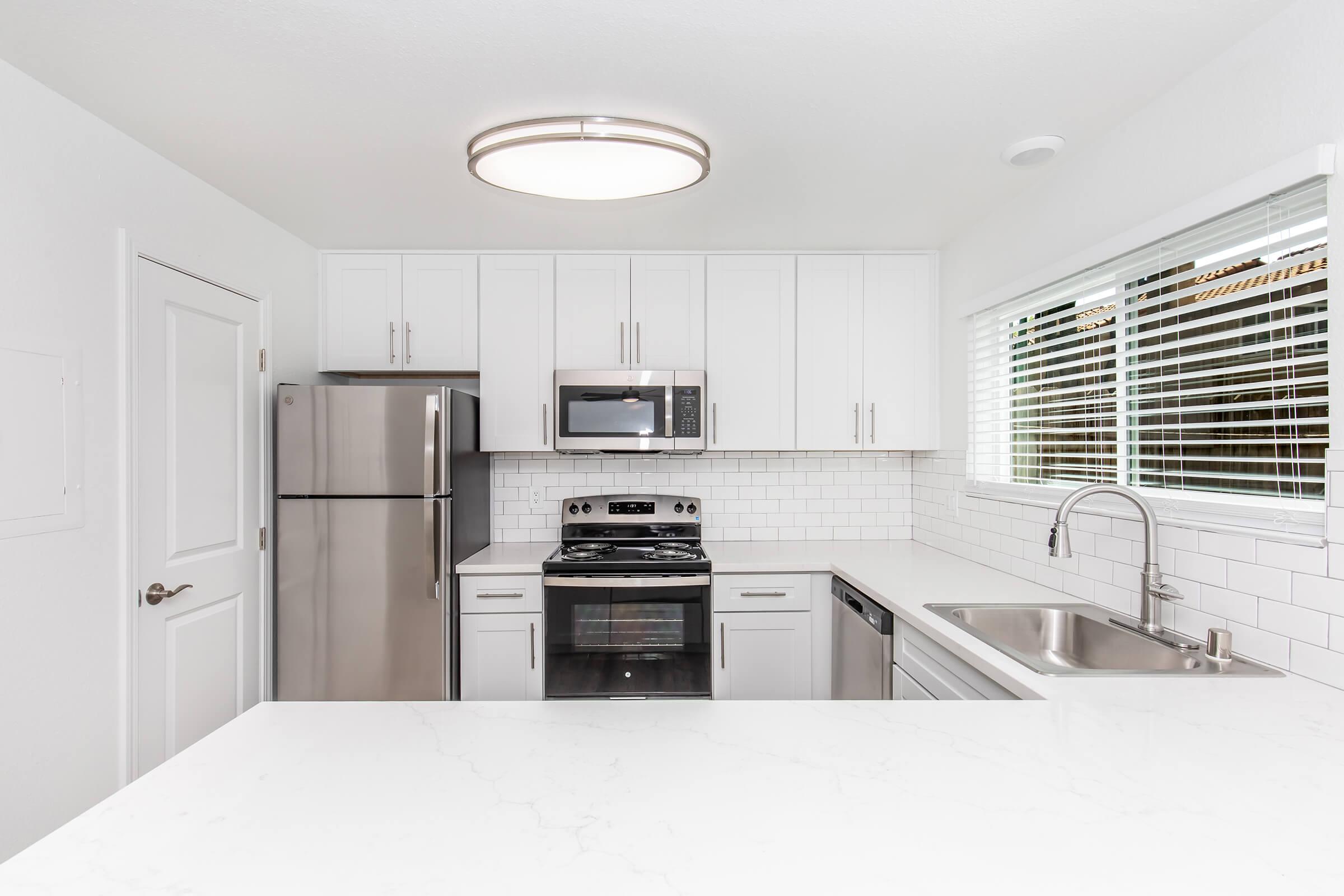 Modern kitchen featuring white cabinetry, stainless steel appliances including a refrigerator, microwave, and oven, a sleek marble countertop, and a double sink. Bright light fixture overhead and large window with blinds allow natural light to enhance the space.