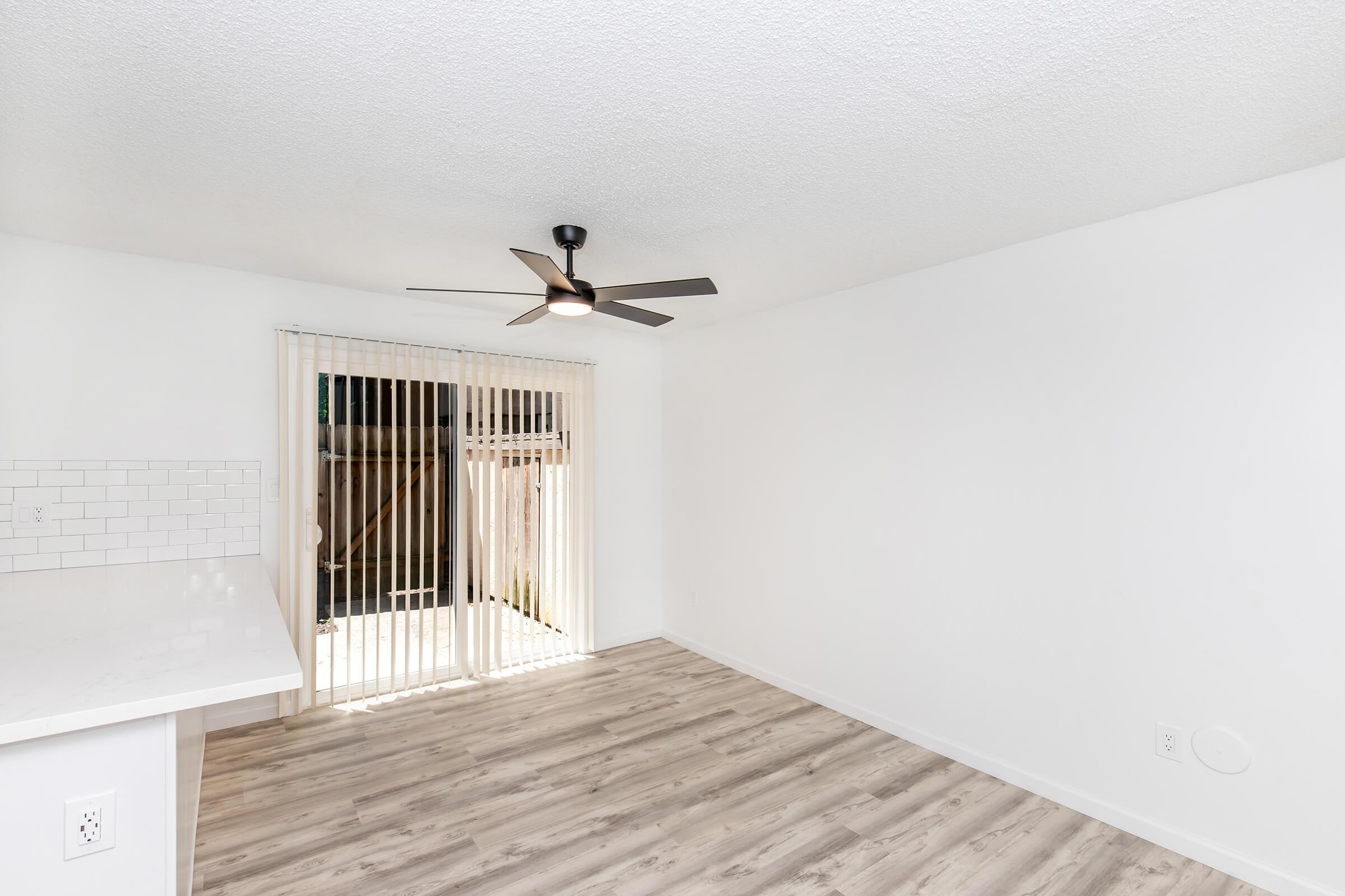 A spacious, bright room with light-colored walls and wooden flooring. It features a ceiling fan and a sliding glass door with vertical blinds leading outside. The room has a minimalist design, creating an open and airy atmosphere.