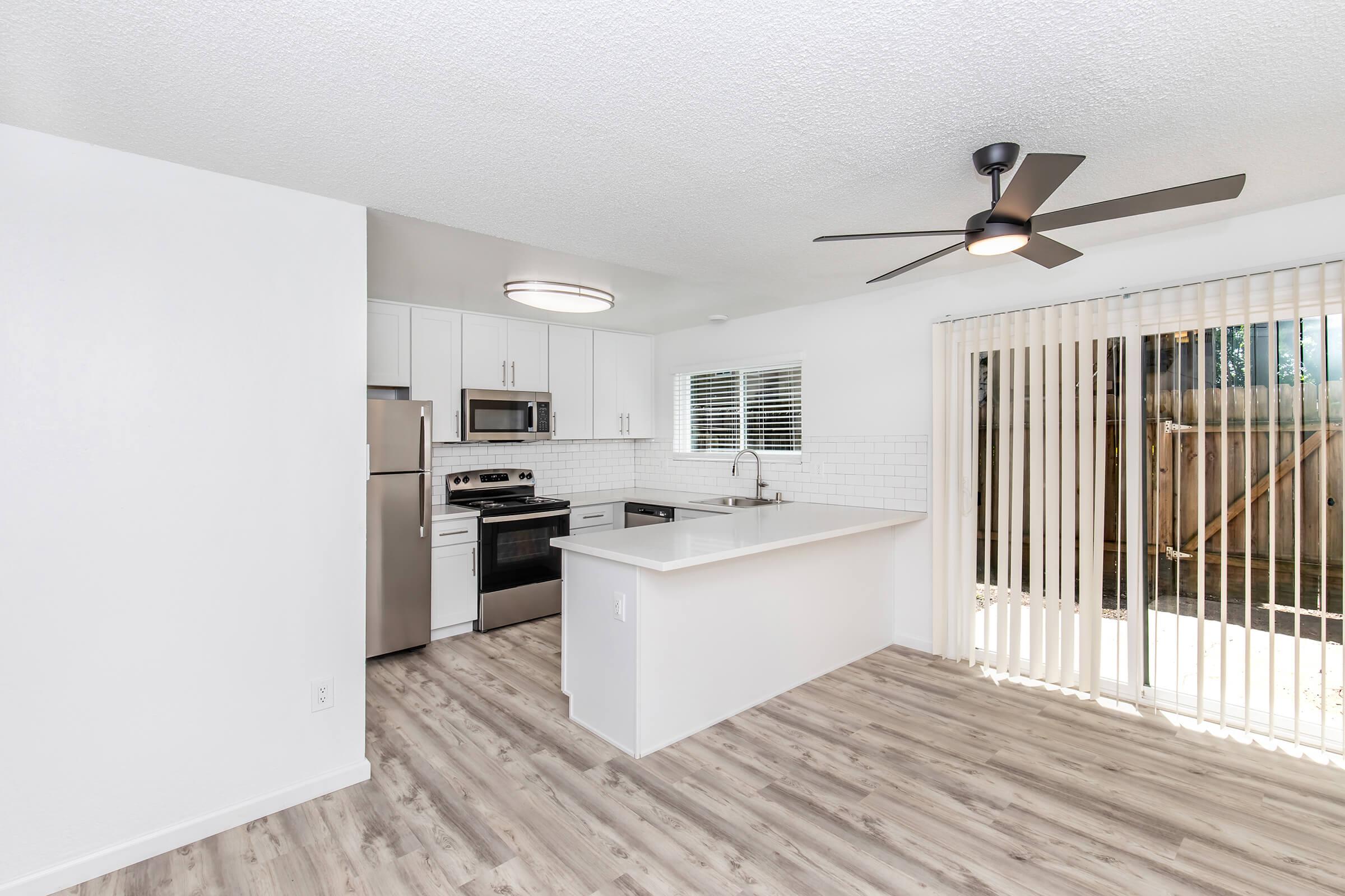 A modern kitchen with stainless steel appliances, including a refrigerator, oven, and microwave. The space features white cabinetry, a large island with a sink, and a ceiling fan. There are sliding glass doors leading to a patio, with light-colored wood-style flooring throughout. Bright natural light fills the area.