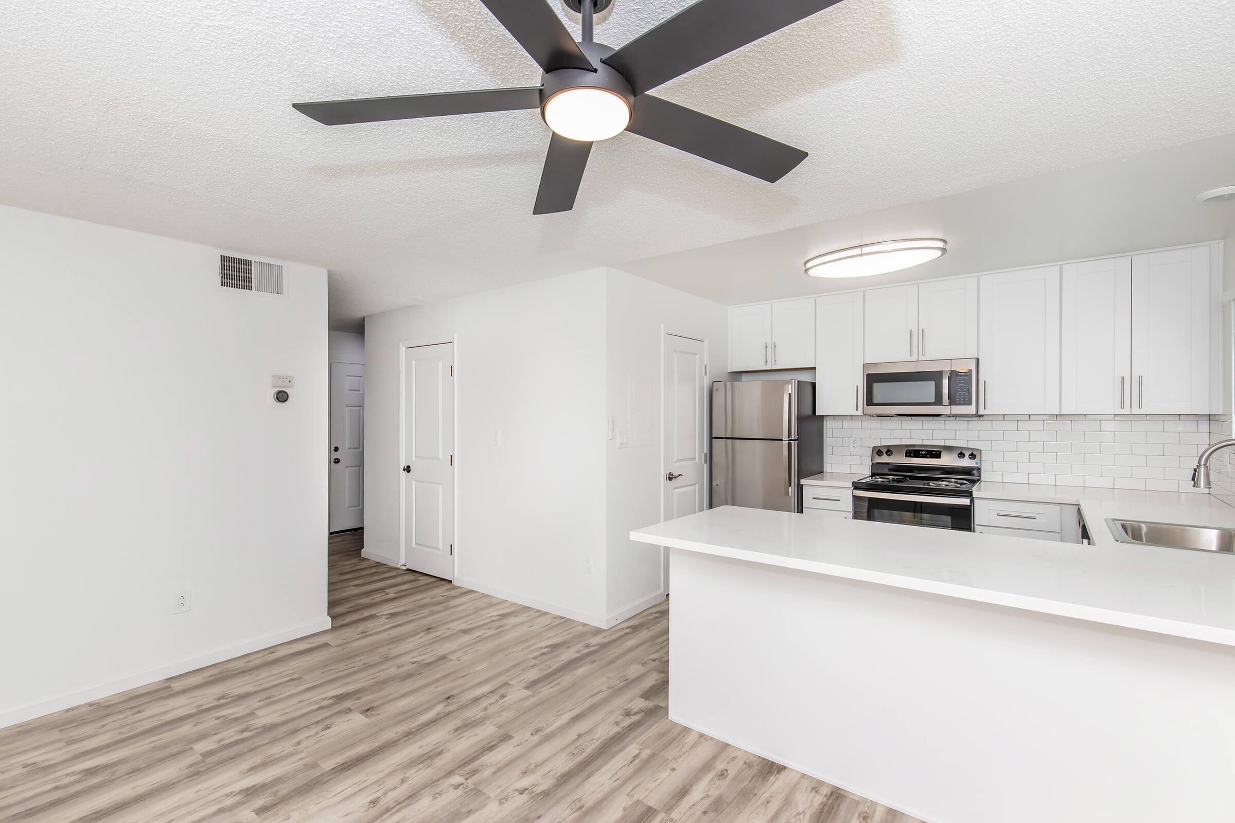 Modern kitchen and living area showcasing white cabinetry, stainless steel appliances, and a sleek island with a countertop. A ceiling fan is visible, and the floor features light wood-like laminate. The walls are painted white, contributing to a bright and open atmosphere.