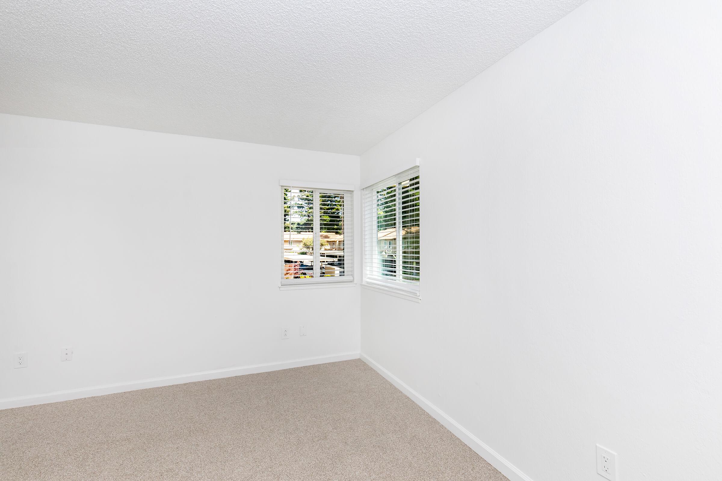 A bright, empty room with white walls and carpet. There are two windows with white blinds allowing natural light to fill the space. The room has a clean, minimalistic design, ready for furnishings or decorations.