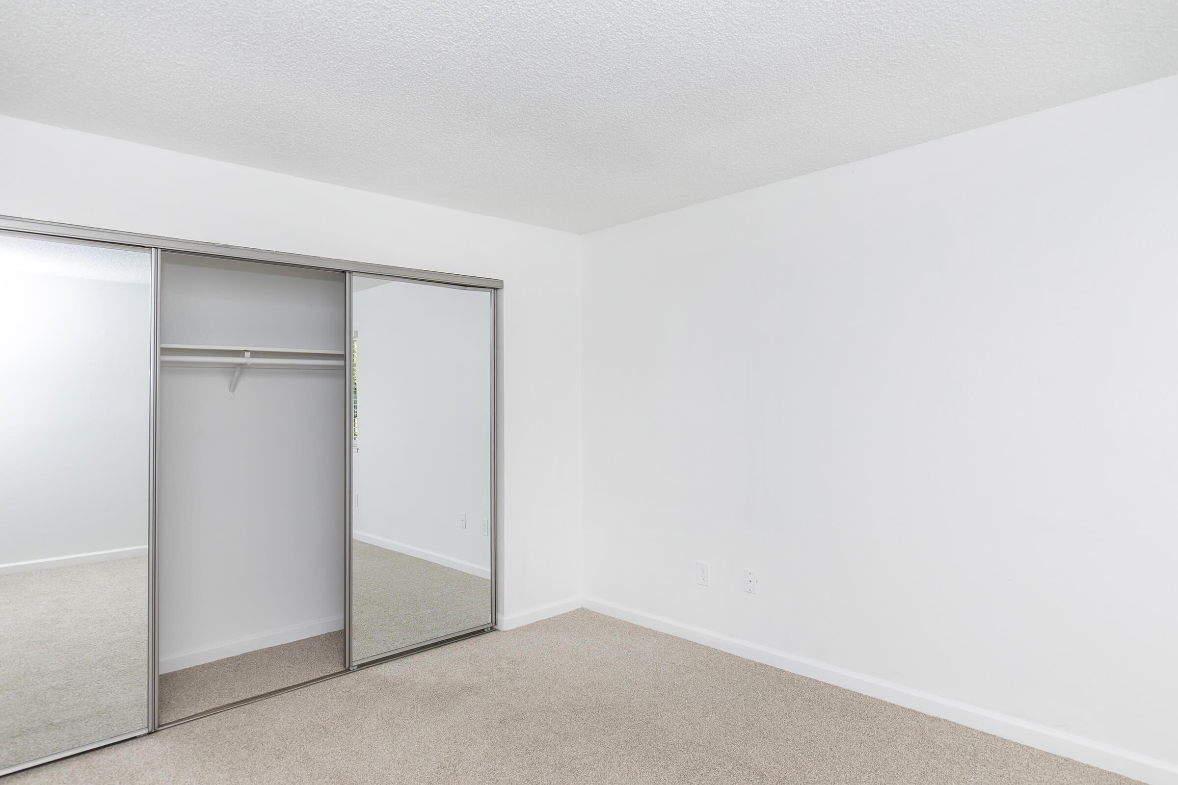 Empty room with a light-colored carpet and white walls. There is a mirrored sliding closet door on one side, showcasing a simple closet design with a hanging rod. The space is well-lit and minimalistic, creating a clean and open atmosphere.
