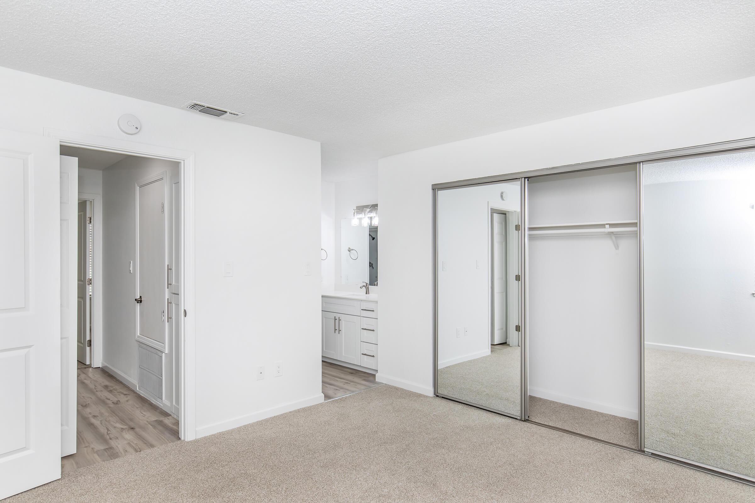A spacious, well-lit bedroom featuring beige carpet flooring. On one side, there is a large mirrored closet with sliding doors, while to the left is a doorway leading to a bathroom with modern fixtures. The walls are painted white, creating a bright and open atmosphere.