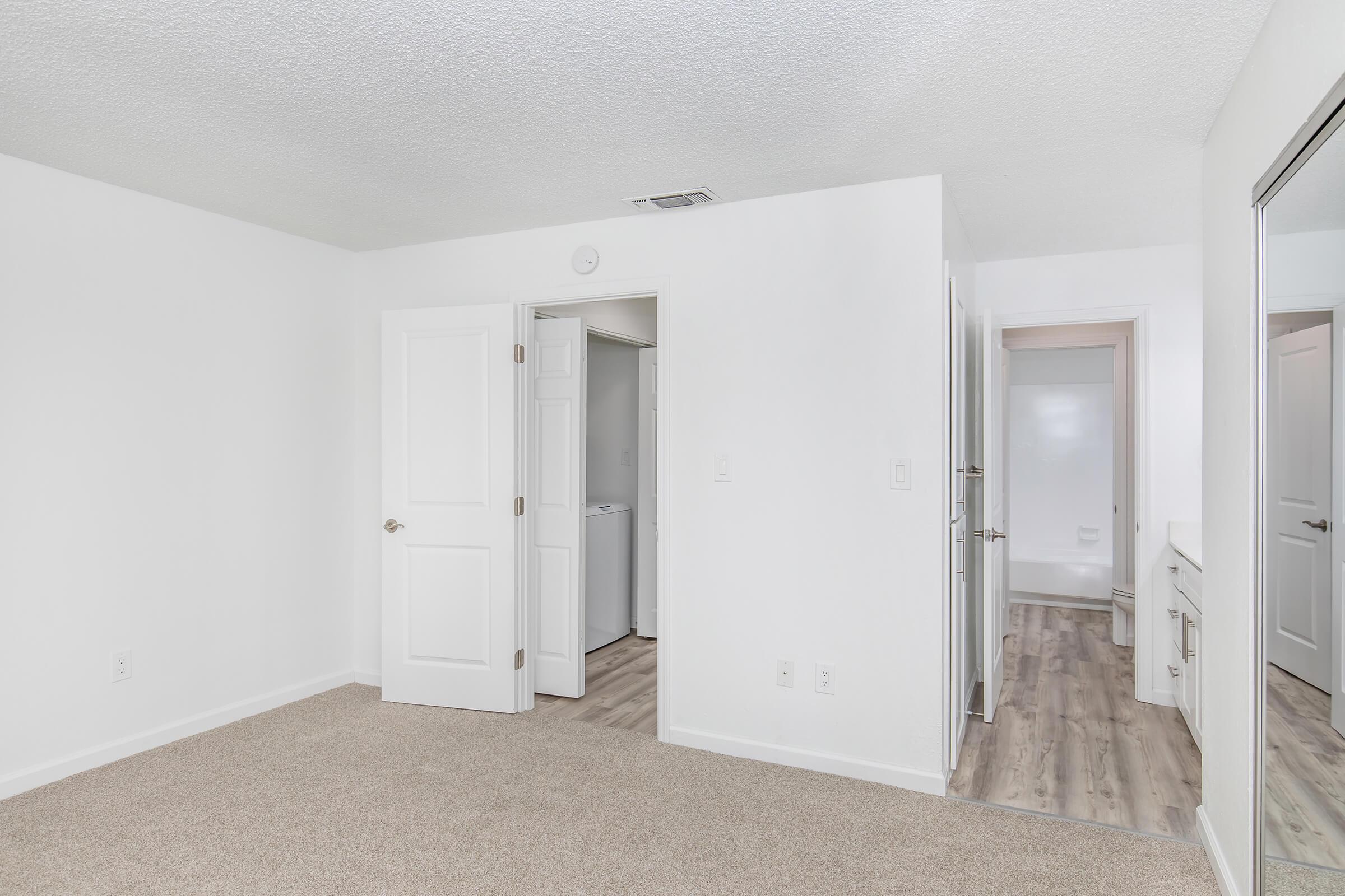 A spacious, empty room with light-colored walls and carpeted flooring. A doorway leads to a bathroom, while another leads to a hallway. Natural light illuminates the space, creating a bright and airy atmosphere. There is a mirror on one wall, and the overall decor is simple and clean.