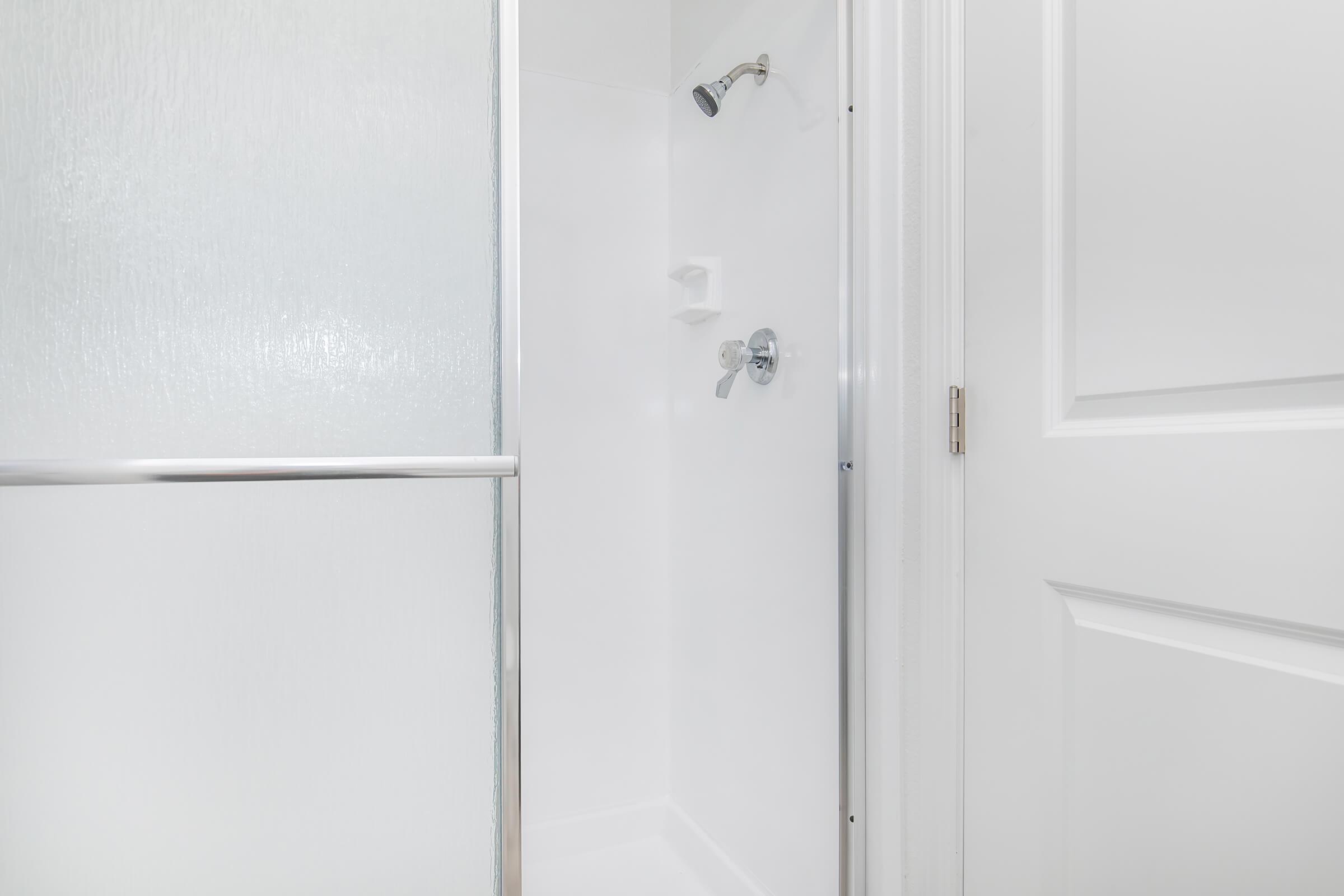 A modern shower stall with a clear glass door. The interior features a simple white wall, a showerhead mounted on the wall, and a control knob for adjusting the water temperature. The space is clean and minimalistic, with bright lighting and a white door leading to or from the bathroom.