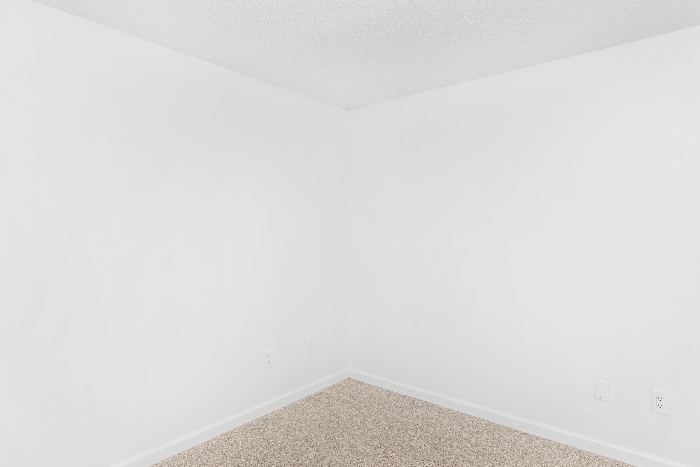 A bare, empty room with light beige carpet and white walls. The space features a clean corner with no furniture or decorations, providing a minimalist and spacious feel.