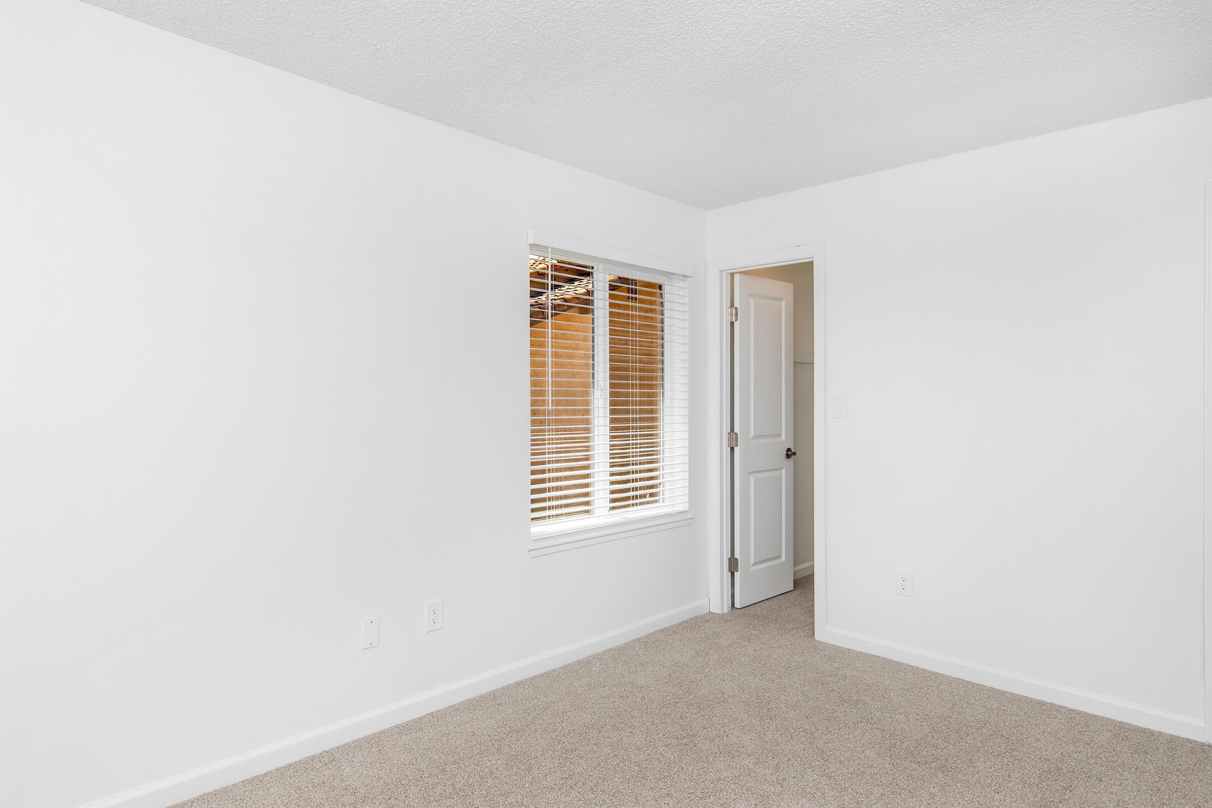 A spacious, empty room with white walls and light beige carpet. There is a window with blinds on the left side, allowing natural light to enter. A white door is visible on the right side, leading to another area. The room has a clean and minimalistic appearance, ideal for various uses.