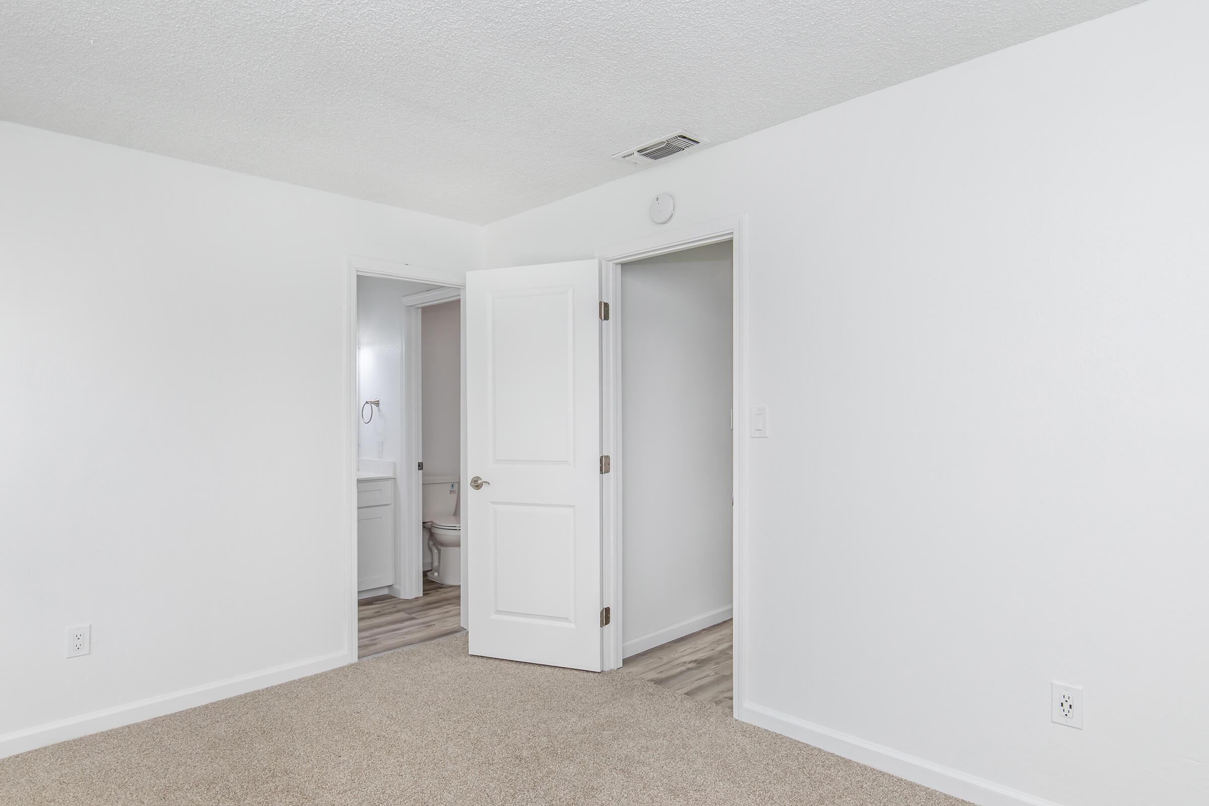 A bright and empty room featuring light-colored walls and carpeted flooring. Two doors are visible: one leading to a bathroom and the other to an adjacent area. The space is well-lit, and there are no furnishings or decorations present, emphasizing a clean and minimalist look.