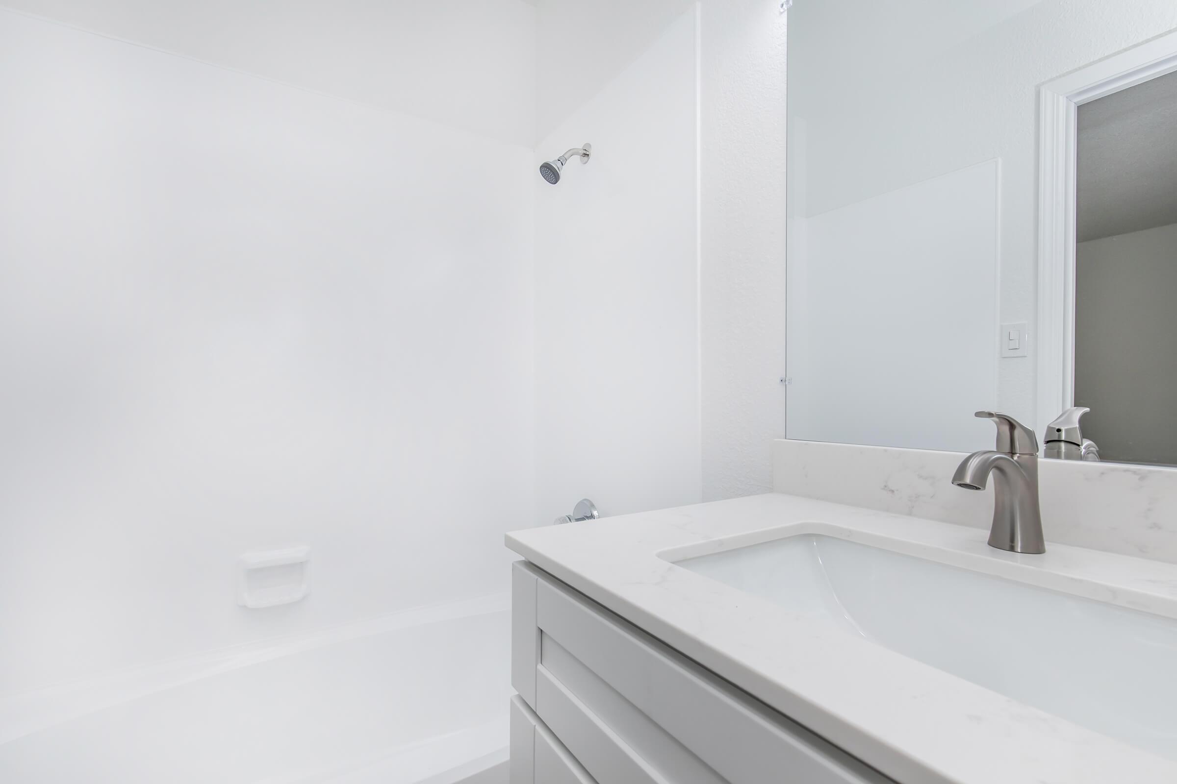 A clean, modern bathroom featuring a white bathtub, a sleek vanity with a light-colored countertop, and a wall-mounted mirror. The walls are plain white, and the shower area includes a simple showerhead. The space appears bright and well-lit, conveying a fresh and airy atmosphere.
