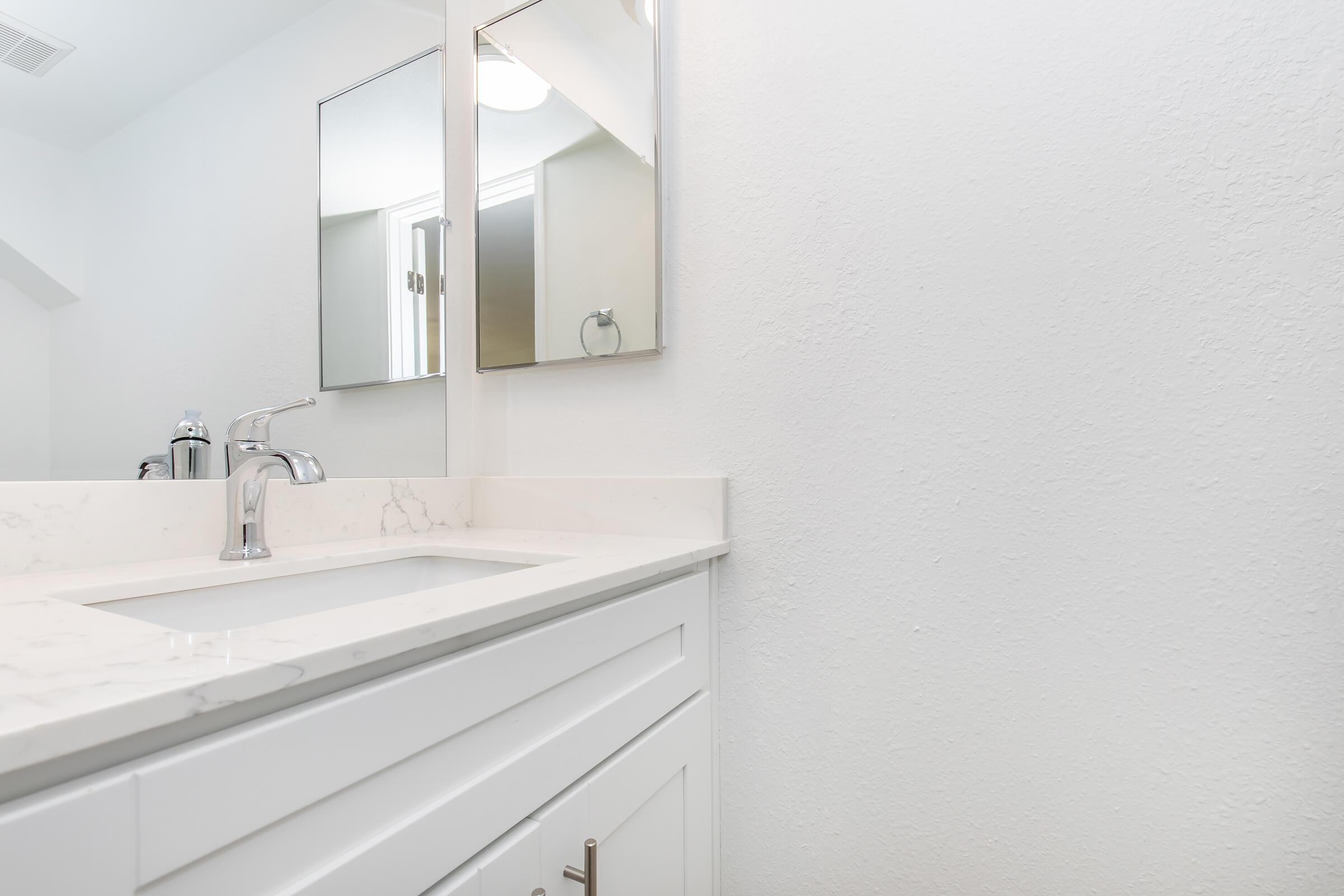 A clean, modern bathroom vanity featuring a white countertop, a sleek faucet, and two mirrors on the wall above. The walls are painted white, creating a bright and spacious atmosphere, while the vanity cabinetry is minimalistic in design.