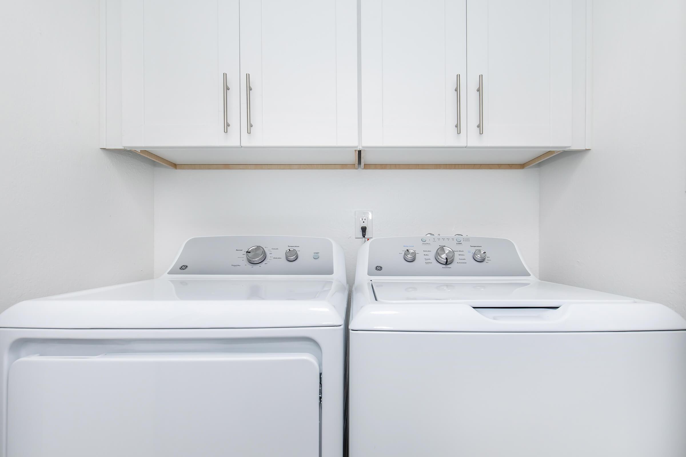 A clean laundry room featuring a white washing machine and dryer side by side, with a minimalistic design. Above them are simple, modern cabinets, providing additional storage space. The walls are painted white, creating a bright and airy atmosphere.