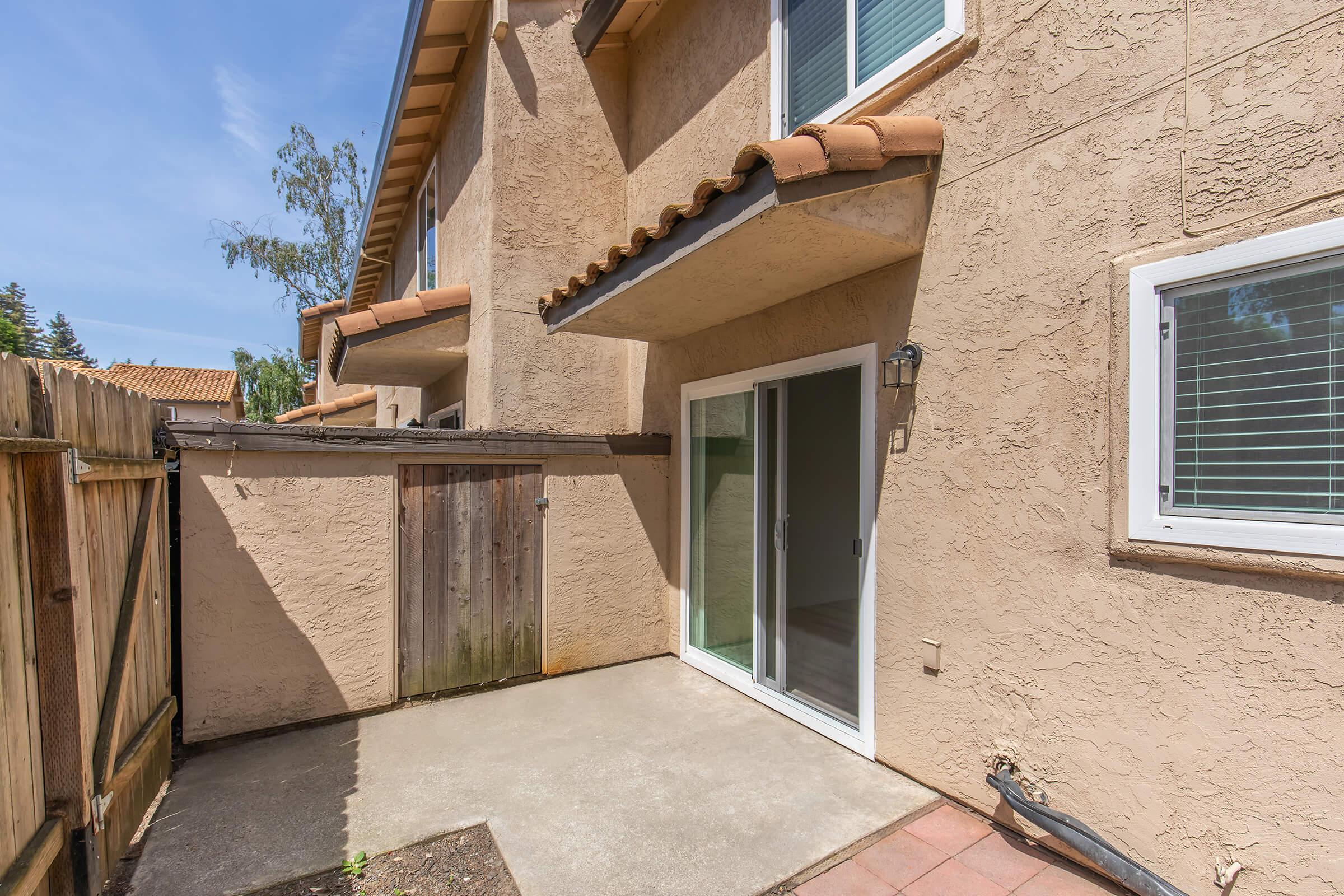 A small outdoor patio space with a concrete floor, featuring a wooden fence on one side and sliding glass doors leading into a house. The wall is textured and appears to be a light beige color, and there are some plants near the edge of the patio. Bright blue sky is visible above.