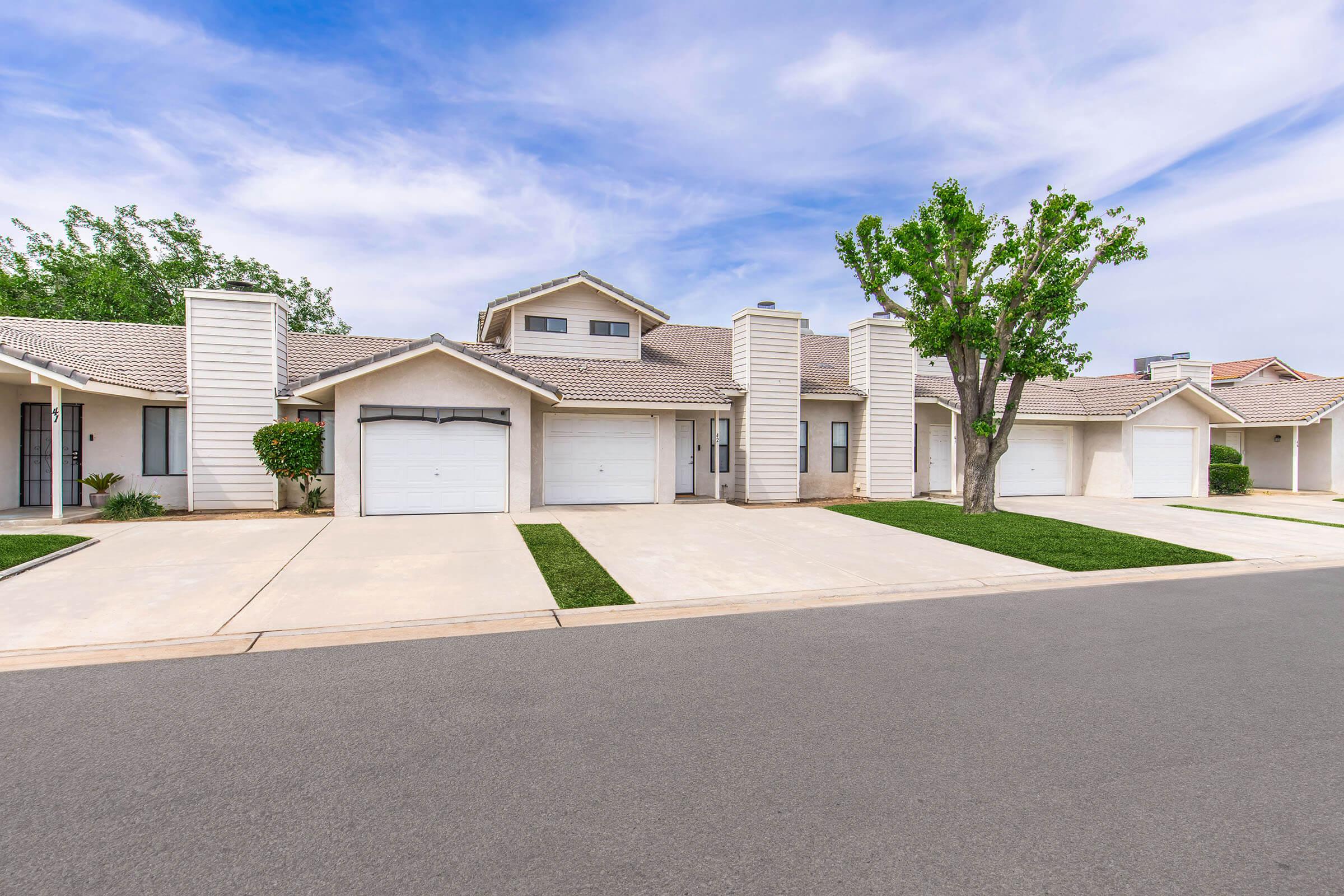 A residential neighborhood featuring a single-story home with a light-colored facade. The property has a driveway leading to a garage, with well-maintained grass and trees in the front yard. The sky is partly cloudy, creating a bright atmosphere.