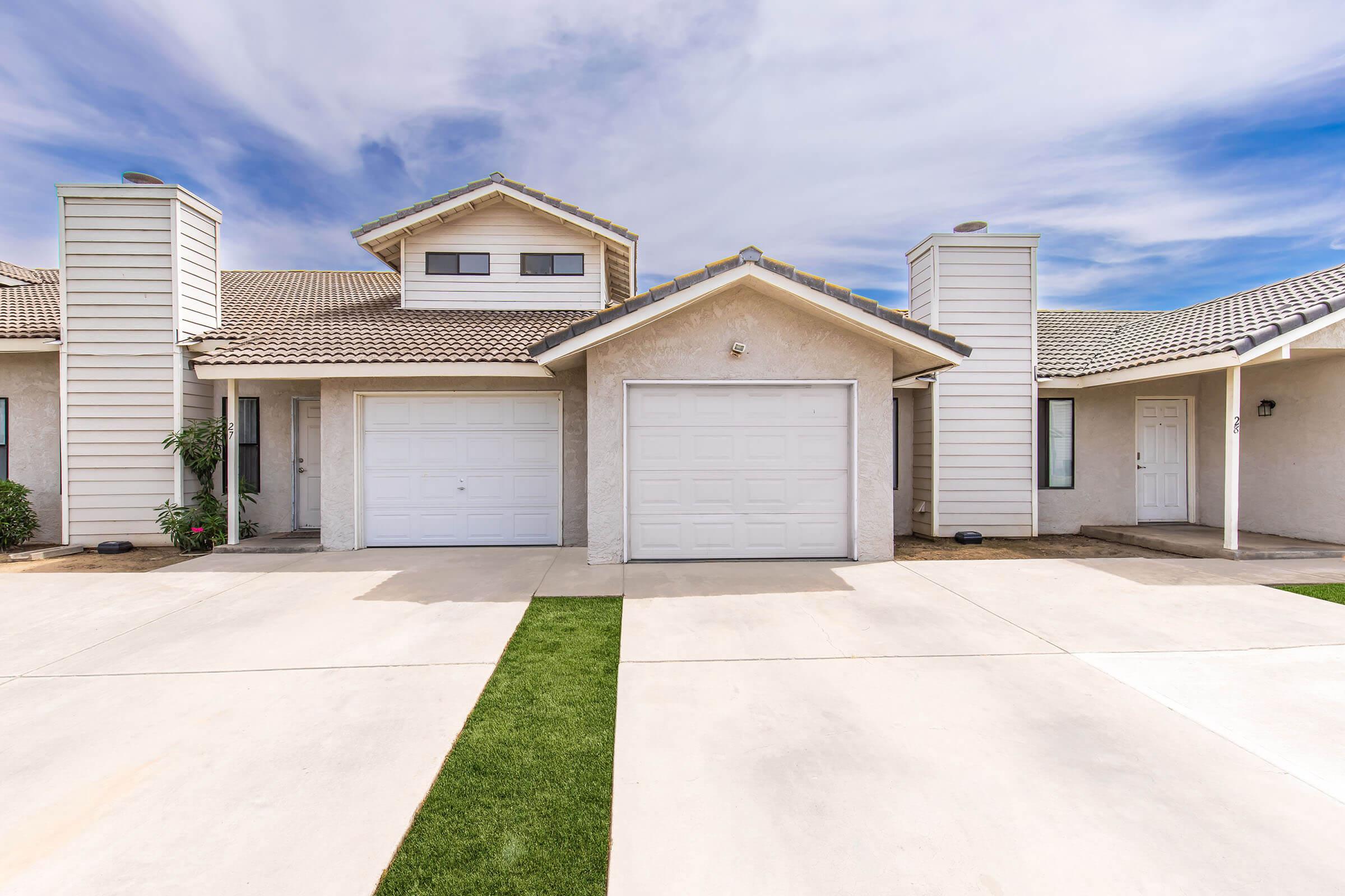 A modern, single-story building with two attached garage doors, a triangular roof, and two decorative towers. The façade is light-colored with a clean concrete driveway leading to green grass patches on either side. The sky is partly cloudy, creating a bright ambiance.