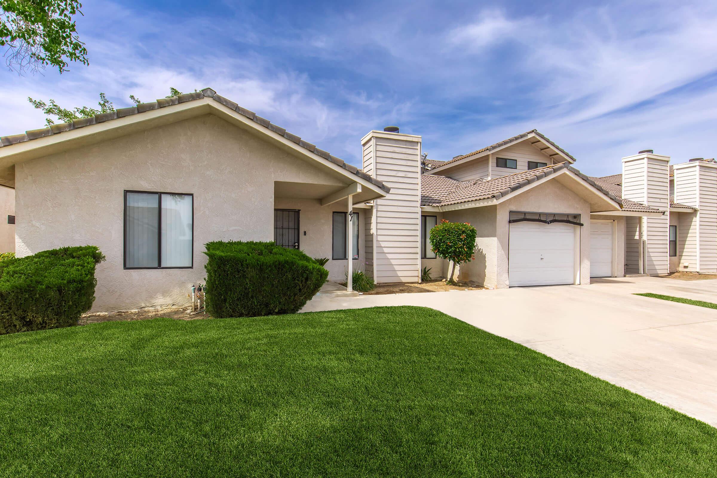 A residential home with a light-colored exterior, featuring a landscaped front yard and green grass. The house has a garage with a door on the right, large windows, and a covered entryway. The sky is partly cloudy, providing a bright and inviting atmosphere.