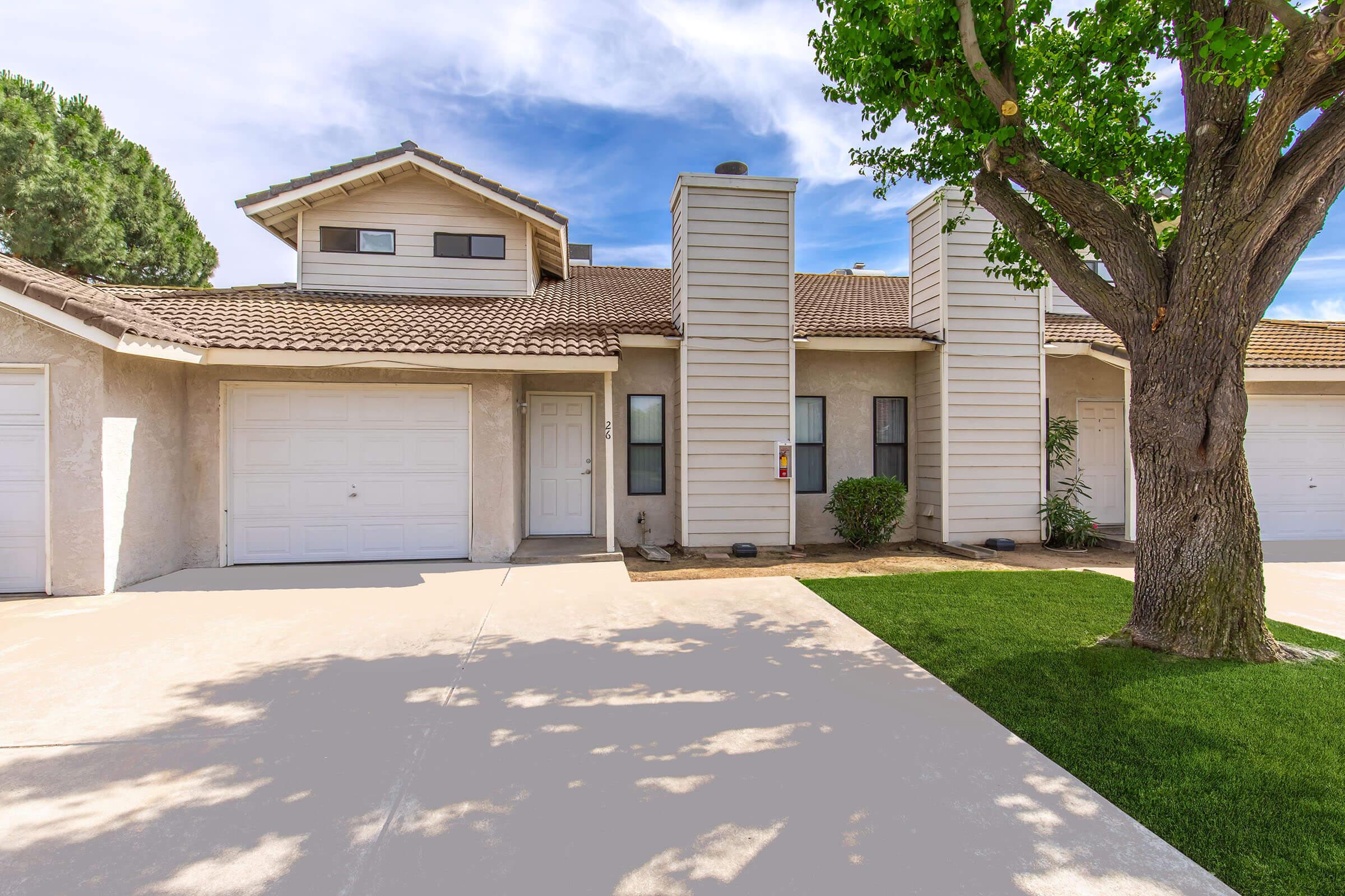 A well-maintained two-story building with a light-colored exterior and a tiled roof. It features two garage doors, multiple windows, and a small landscaped yard with green grass and a tree. The sky is clear with some clouds, creating a bright and inviting atmosphere.