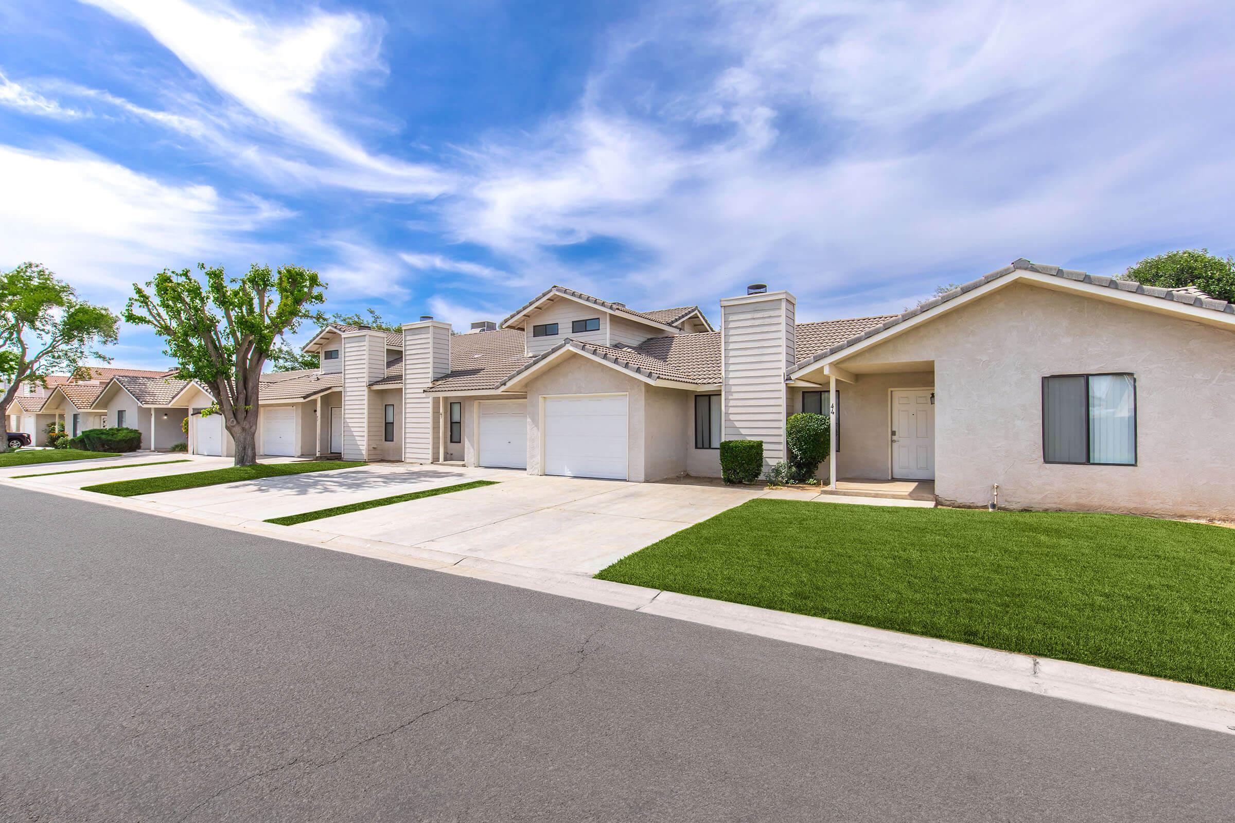 View of a suburban street featuring a row of houses with one-car garages. The homes have a neutral color palette and manicured lawns. A clear blue sky with scattered clouds complements the scene, creating a serene residential atmosphere. The street appears well-maintained and inviting.