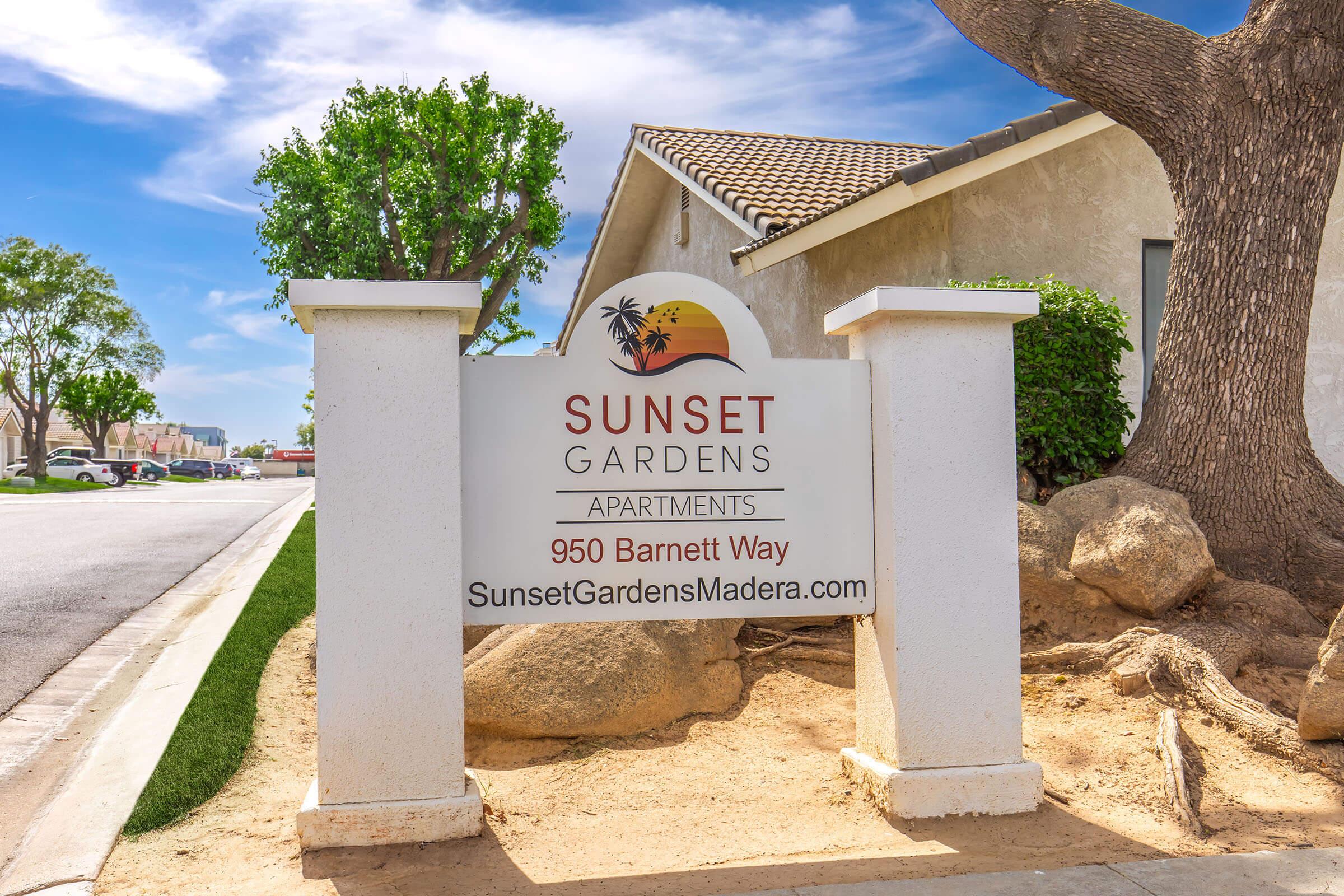 Sign for Sunset Gardens Apartments, featuring the name "Sunset Gardens" with a sun and palm tree graphic. The address listed is 950 Barnett Way, and the website is SunsetGardensMadera.com. The sign is located near a landscaped area with trees and rocks.