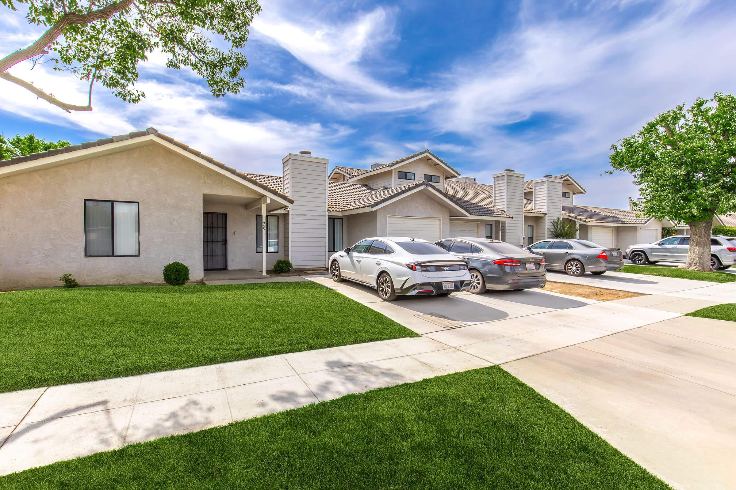 A residential area featuring multiple houses and parked cars. The houses have light-colored exteriors and well-maintained lawns with green grass. The sky is partly cloudy, creating a bright and inviting atmosphere.