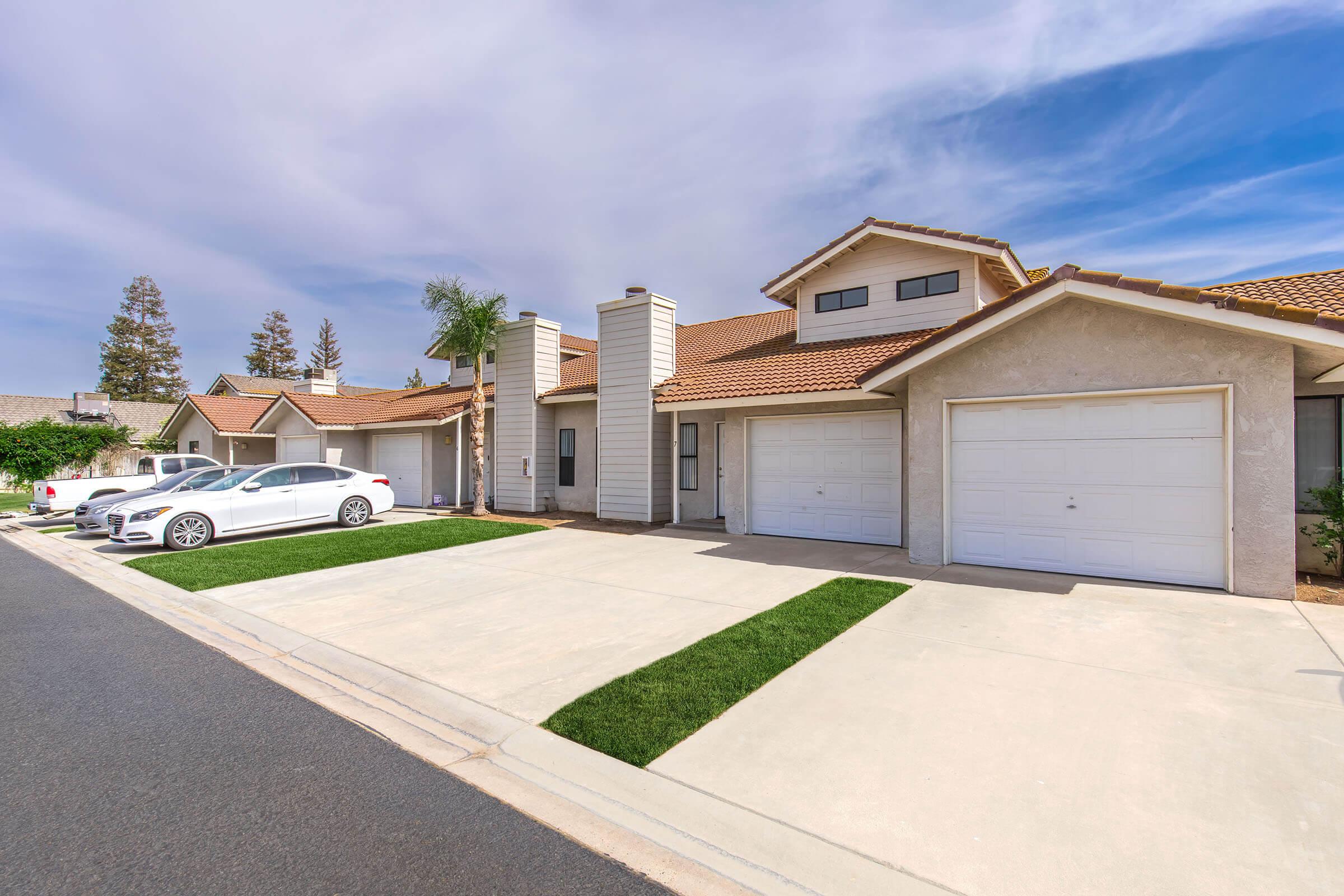A suburban neighborhood showing a row of single-story houses with garages. Three white cars are parked in the driveway, and well-maintained grass patches flank the concrete driveway. The sky is blue with scattered clouds, and there are trees in the background, contributing to the peaceful residential atmosphere.