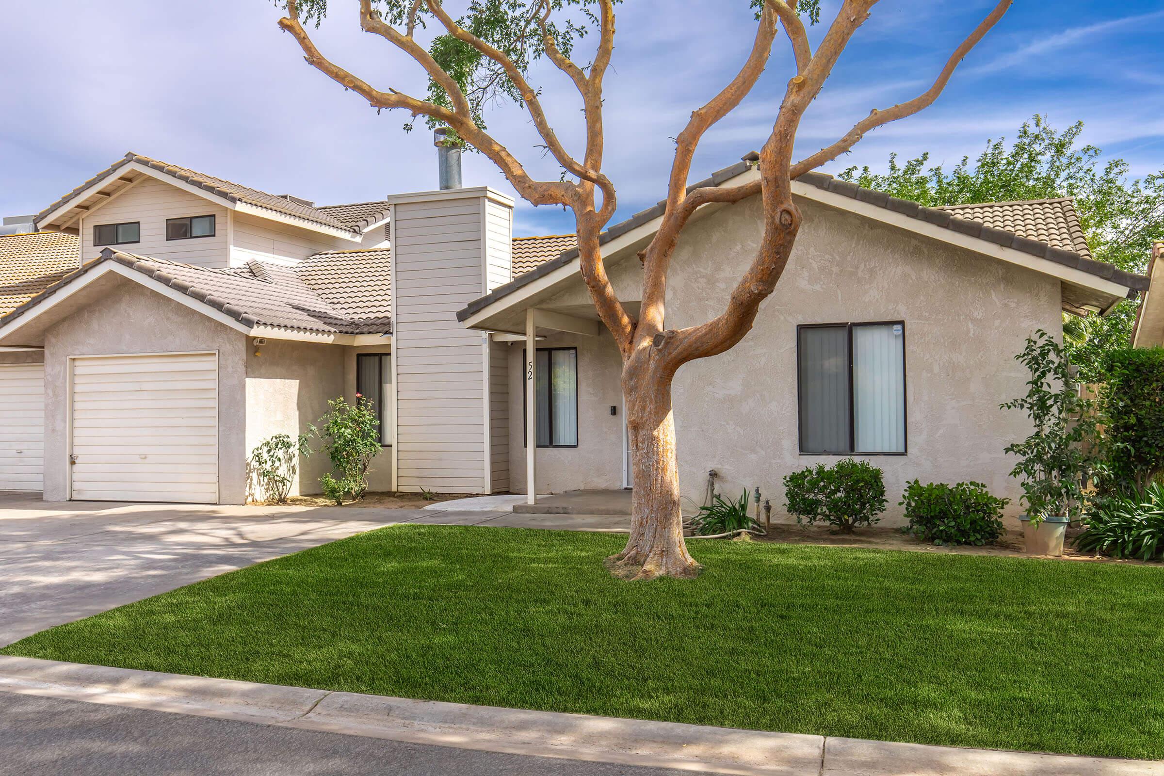 A suburban house with a light-colored exterior, a sloped roof, and a front porch. In the foreground, a manicured lawn with a small tree. The driveway leads to a garage on the left side, and there are various plants and shrubs along the pathway to the entrance. Clear sky in the background.