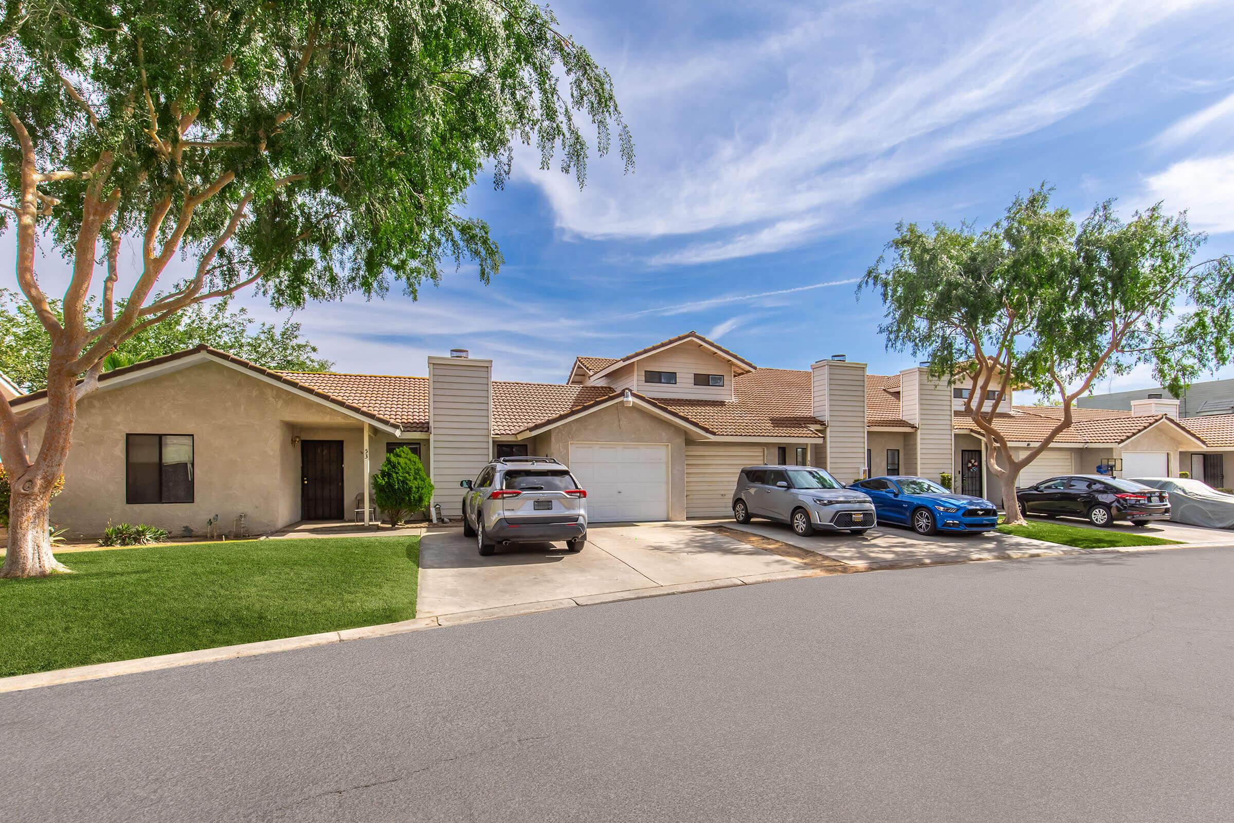 A row of suburban homes with a neatly paved driveway. Three parked cars are visible, along with green trees lining the front yard. The sky is clear with a few clouds, and the homes have light-colored exteriors and sloped roofs. The area appears well-maintained and inviting.