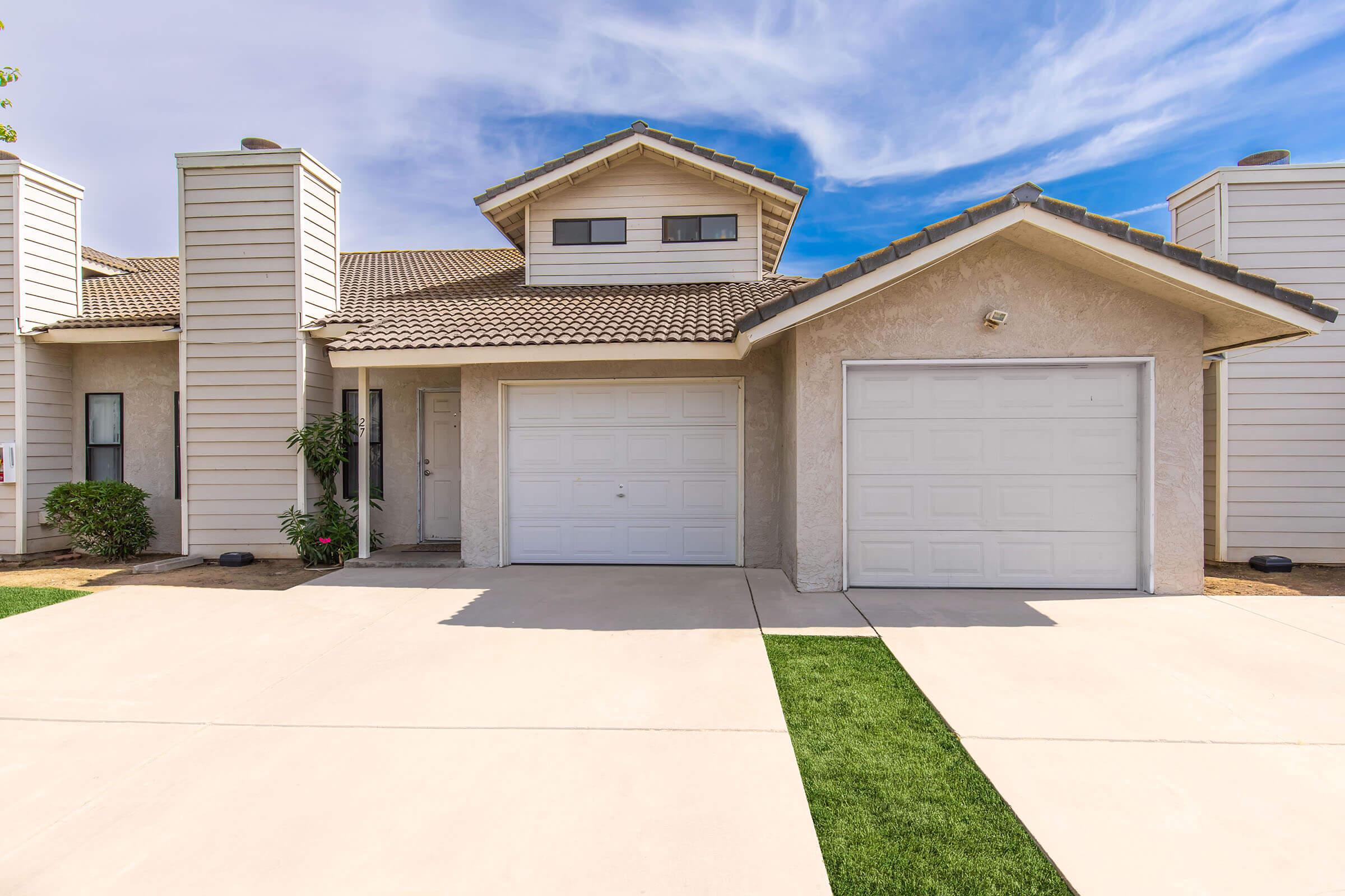 A modern two-story house with a dual garage, featuring a light exterior and a tile roof. The entrance is framed by small shrubs, and a well-maintained lawn with concrete driveway leads up to the property. The sky above is clear with some clouds.