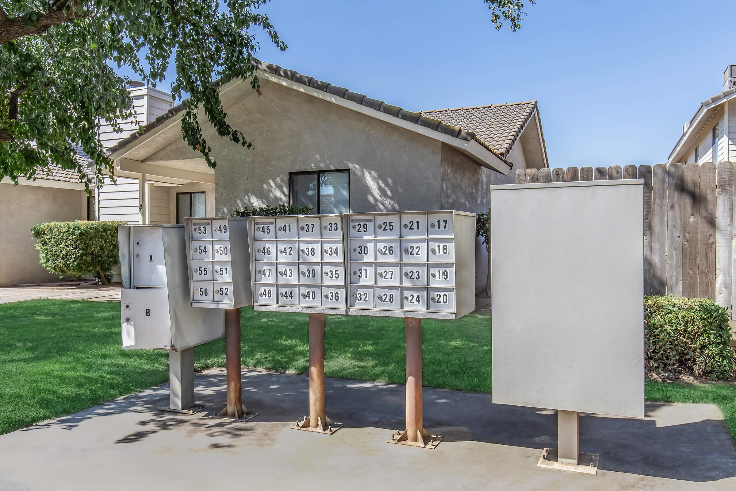A row of mailboxes is situated on a concrete slab in front of a residential house. The mailboxes are organized in a grid pattern, displaying numbers from 1 to 36. A green lawn surrounds the area, and a wooden fence is visible in the background alongside the house with a tiled roof. Clear blue sky is overhead.