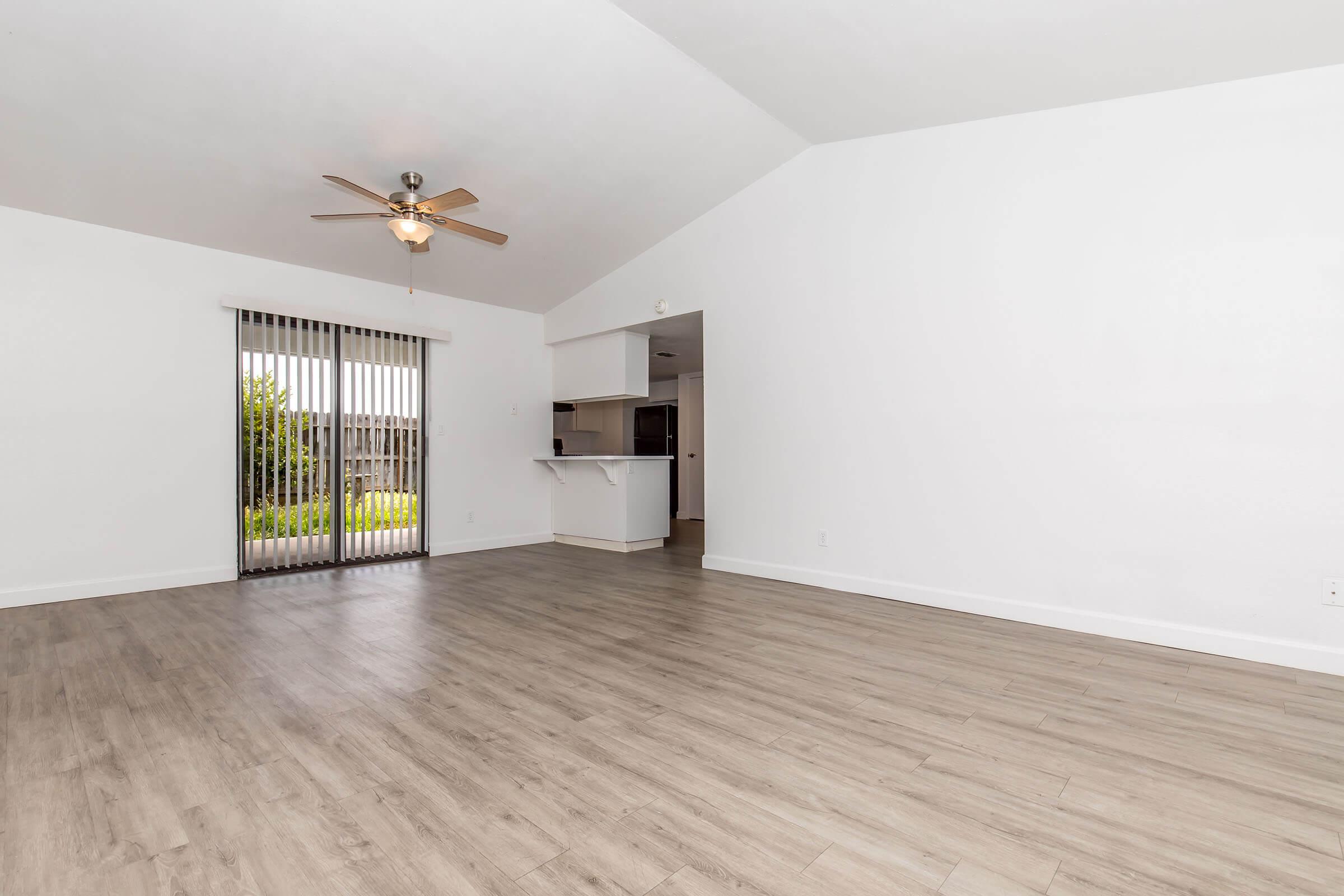 Spacious and bright empty living room with light-colored walls and a high ceiling. Features a ceiling fan, large windows with vertical blinds, and laminate flooring. A doorway leads to an adjoining area, possibly a kitchen. The overall ambiance is open and airy, perfect for personalization.