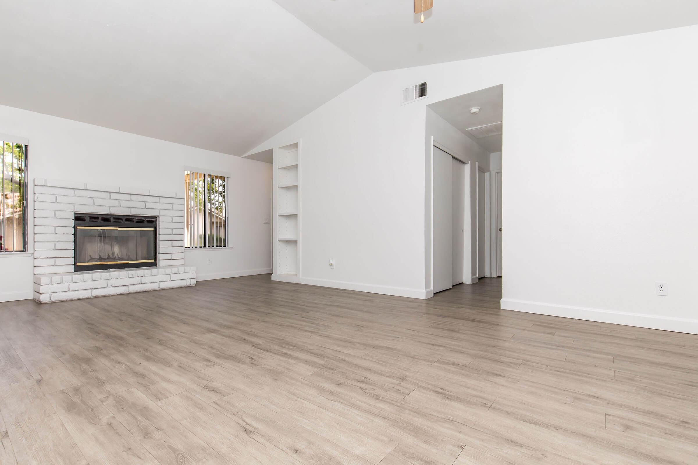 A spacious living room with a high ceiling, featuring a white brick fireplace, large windows with natural light, and light-colored wood flooring. There are built-in shelves on one wall and a doorway leading to another room, creating an open and inviting atmosphere.