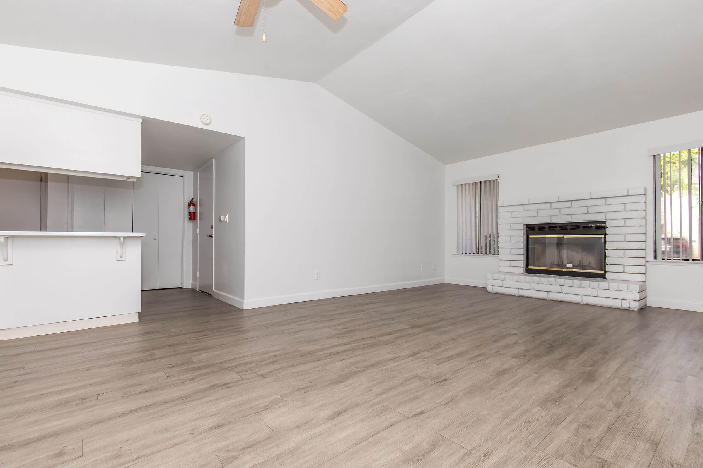 Bright, spacious living room with a high vaulted ceiling and wood laminate flooring. A white brick fireplace is set against one wall, complemented by large windows with vertical blinds. An open layout transitions into a nearby area with a doorway leading to another room. The overall atmosphere is modern and inviting.