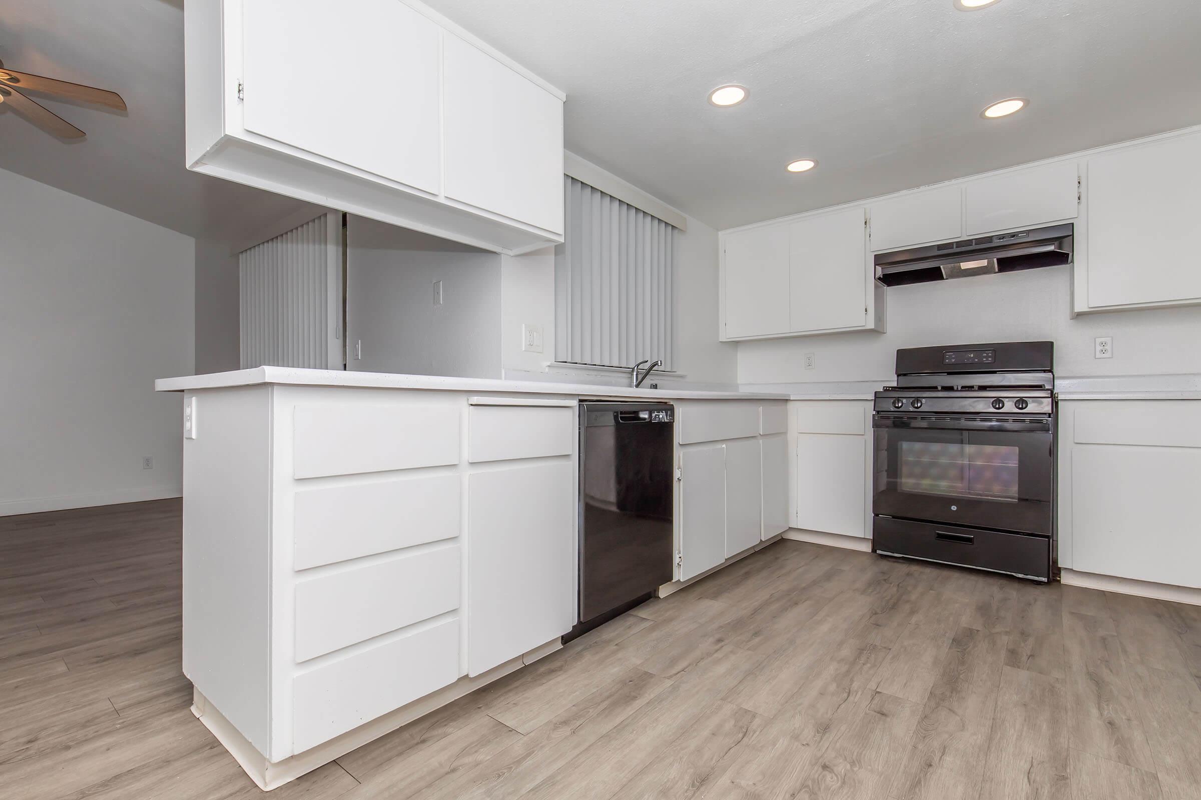 Modern kitchen featuring white cabinetry, stainless steel dishwasher, and black gas stove. The countertop is sleek and light-colored, with an open layout leading to a spacious living area. The flooring is light wood laminate, and there are multiple ceiling lights providing bright illumination.