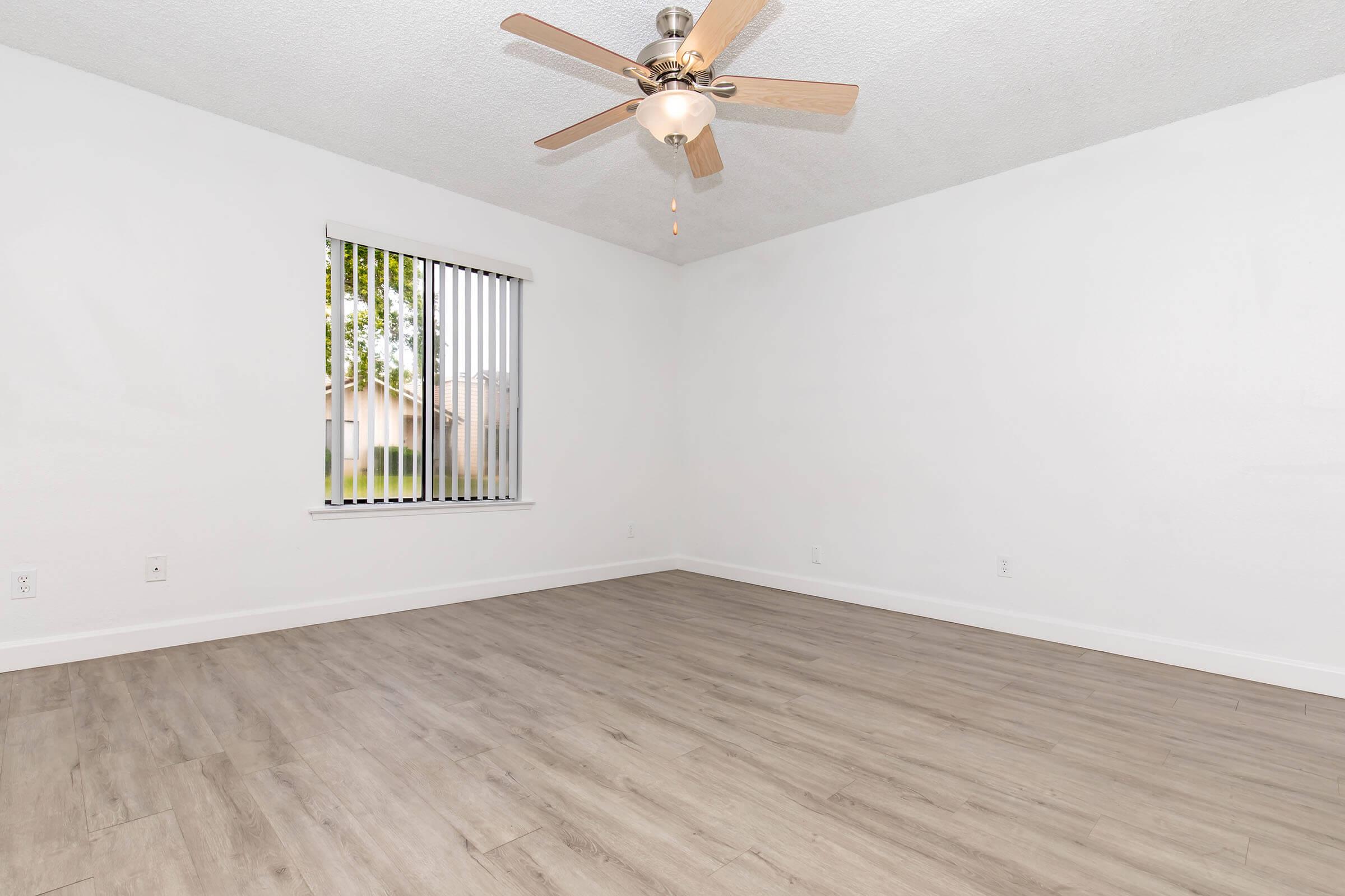 A spacious, empty room with light-colored walls and wood laminate flooring. A ceiling fan with wooden blades is displayed, and a window with vertical blinds allows natural light to enter. The room is well-lit and has a clean, minimalist aesthetic.