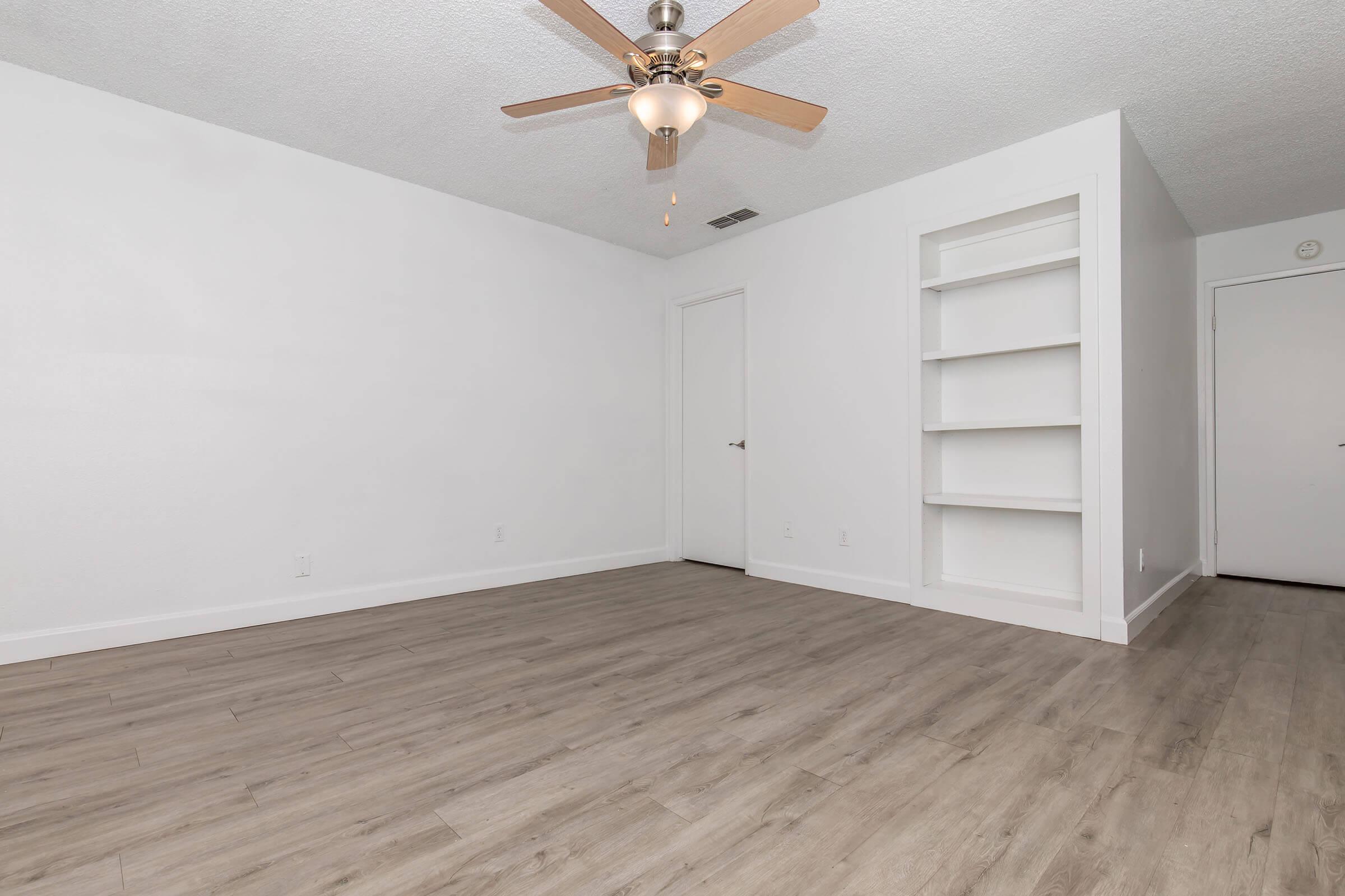 A spacious, empty room featuring light wood flooring, white walls, and a ceiling fan with wooden blades. A small built-in shelf is visible on one side, and a closed door can be seen on the left, providing access to another area. The overall atmosphere is bright and minimalist.