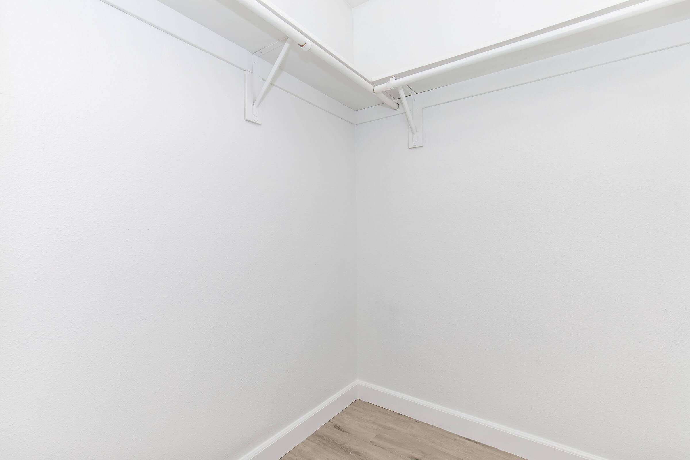Empty corner of a closet with white walls and a light-colored wooden floor. There are two wall-mounted brackets for hanging shelves or rods, but no shelves or clothing are present in the space. The overall atmosphere is bright and minimalistic.
