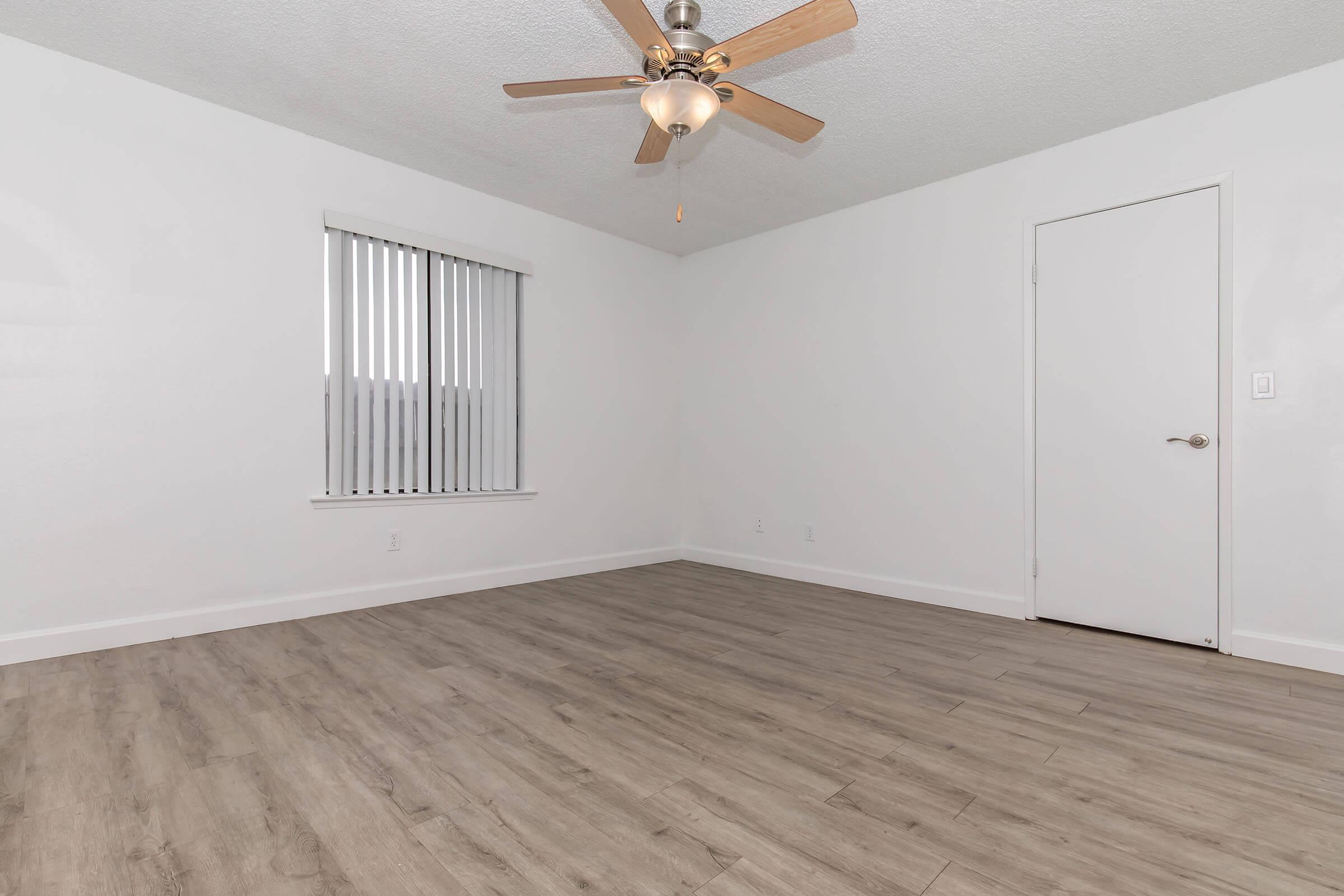 A spacious room with light-colored walls and a wooden floor. There is a ceiling fan with wooden blades, a window with vertical blinds allowing natural light, and a white door. The room is empty, creating an open and airy atmosphere.