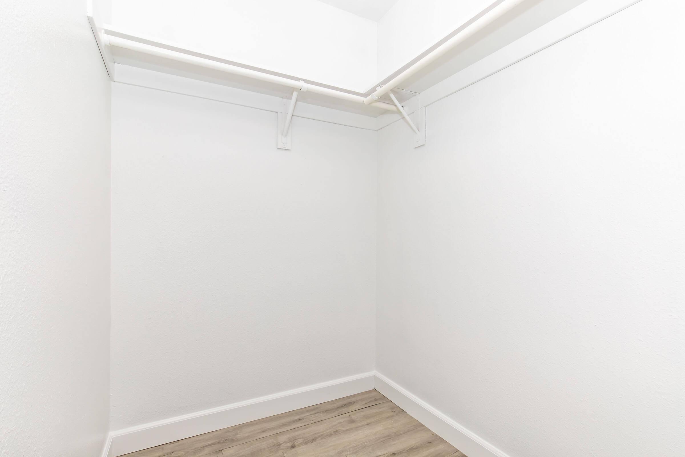 Empty closet with white walls and a wooden floor, featuring two hanging rods for clothes. The space is uncluttered and well-lit, providing a clean, minimalist appearance.