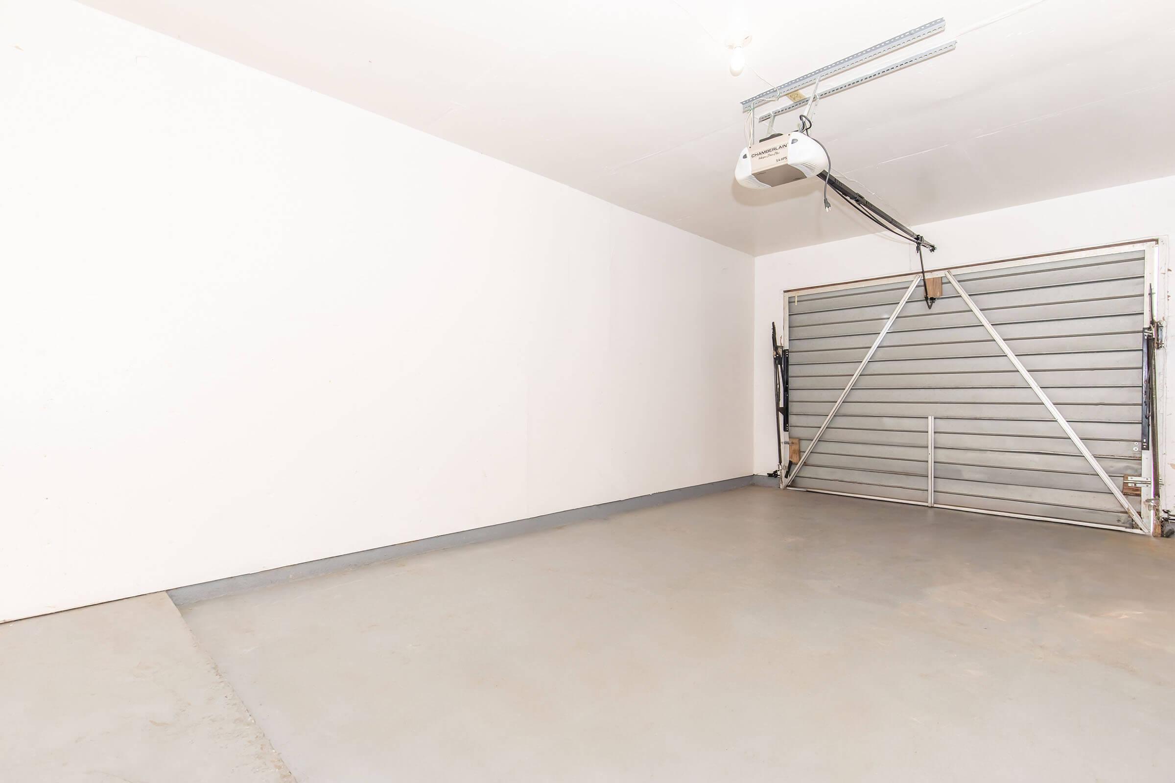 An empty garage with a smooth concrete floor and white walls. The garage door is partially open, revealing a metal garage door with slats. The lighting is bright, providing a well-lit, uncluttered space suitable for storage or parking vehicles.
